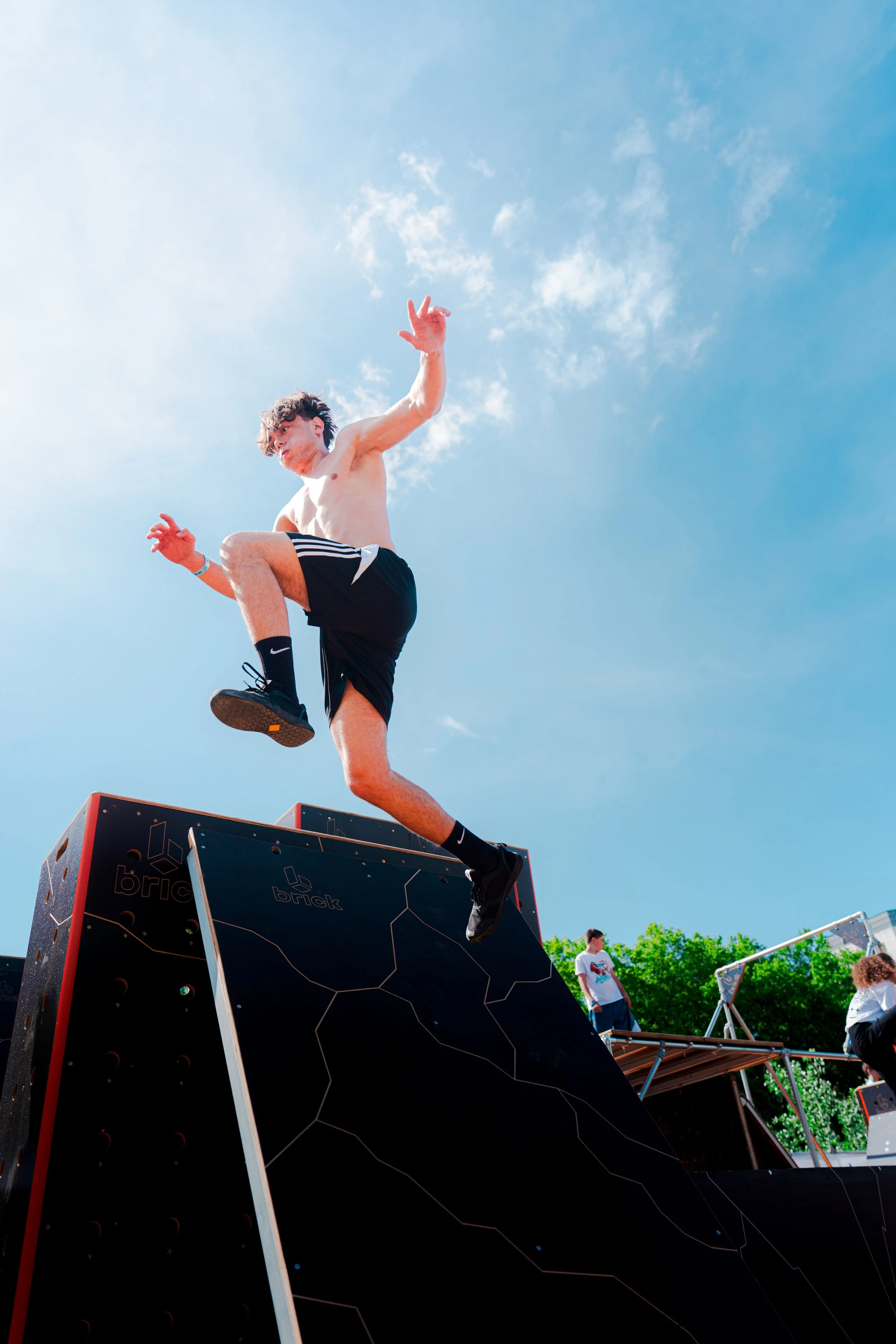 Un jeune homme en short court noir et sans t-shirt saute en l'air depuis une plateforme en formant une position de saut acrobatique, sous un ciel bleu clair avec quelques nuages, lors d'un événement extérieur.