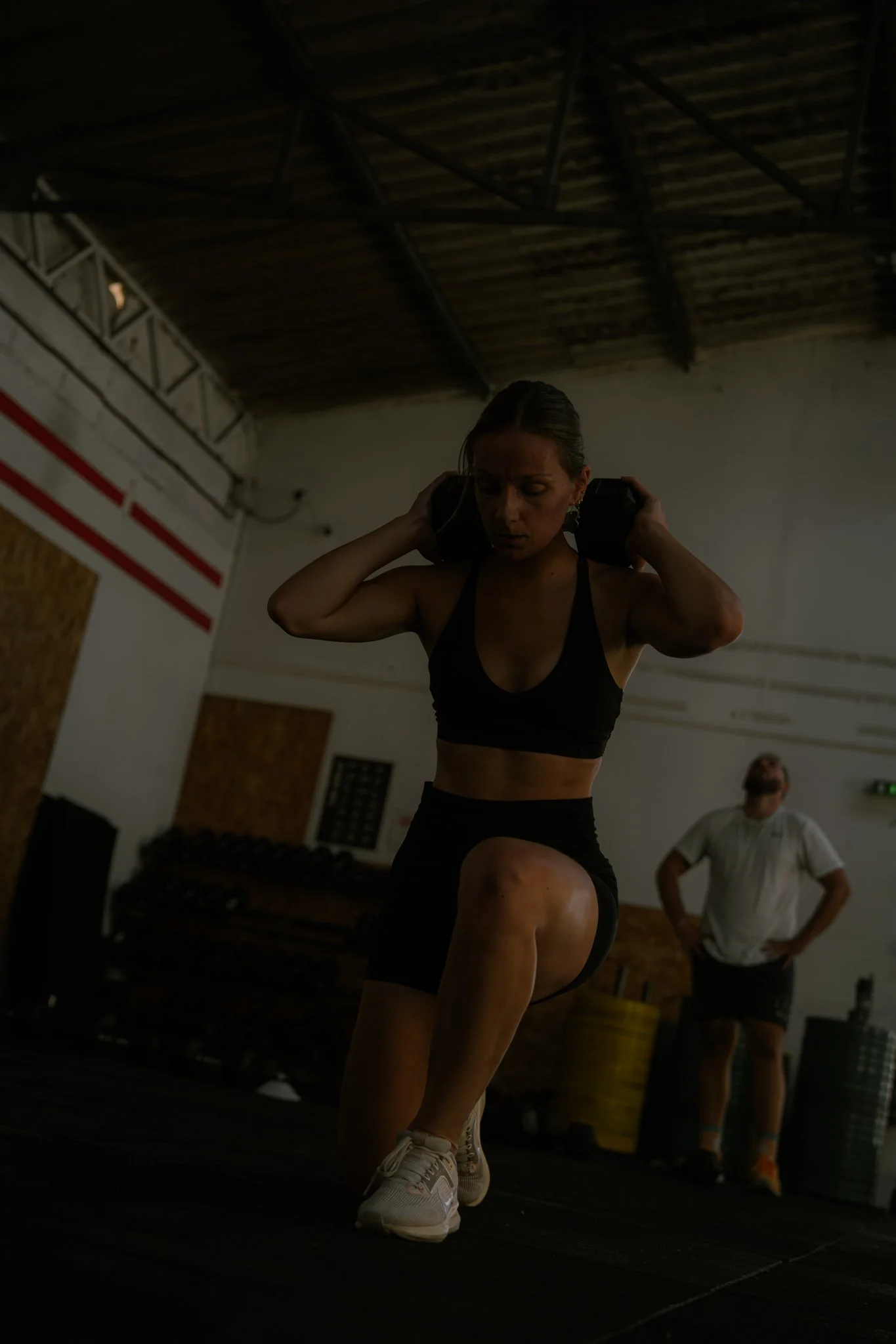 Une femme s'entraîne en salle de sport, un homme en arrière-plan observe. La femme porte un débardeur noir, des shorts et des chaussures de sport blanches. Elle effectue un exercice de musculation sur une surface noire.