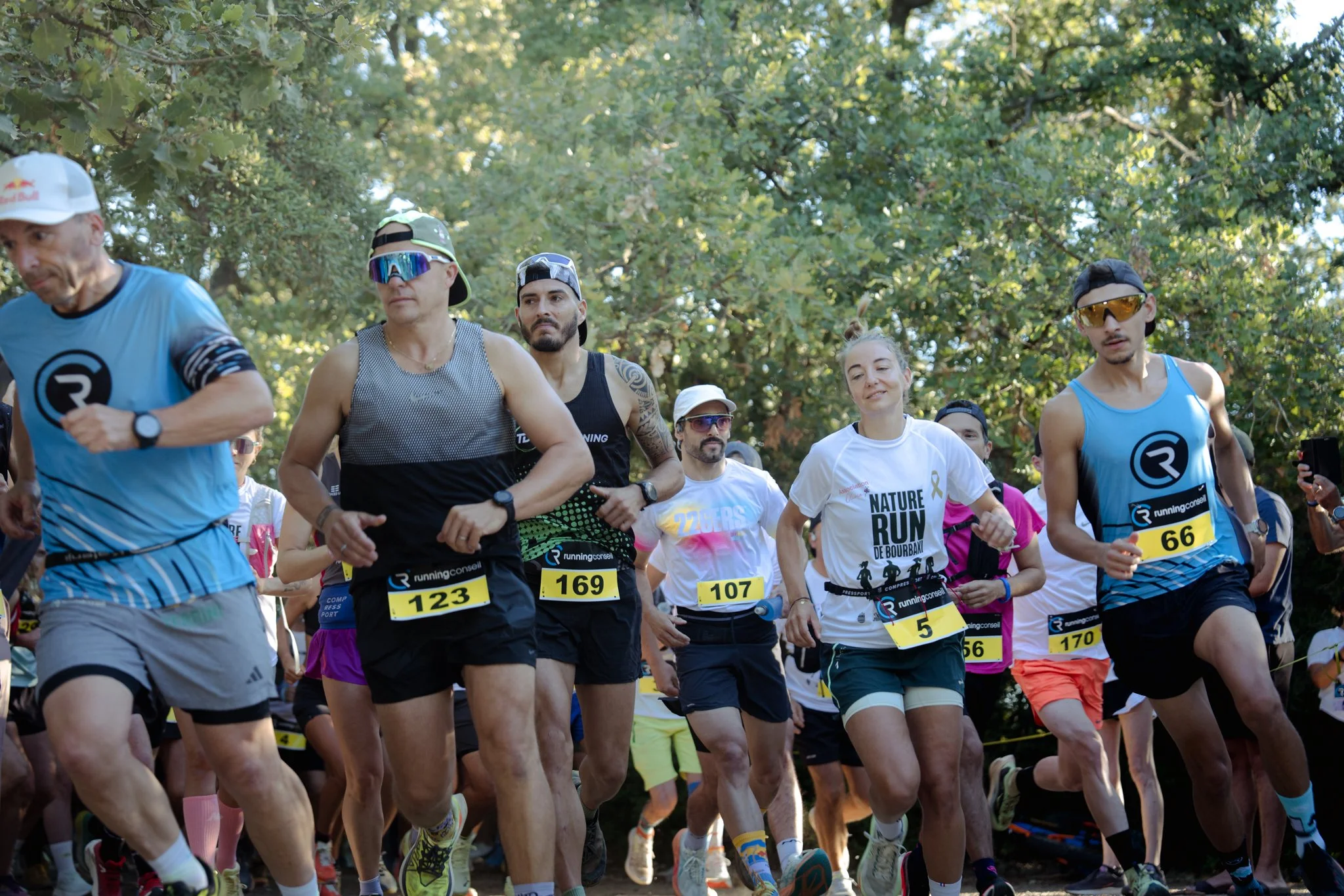 Groupe de coureurs lors d'une course en plein air, portant des t-shirts et des dossards, courant sur un chemin entouré d'arbres.