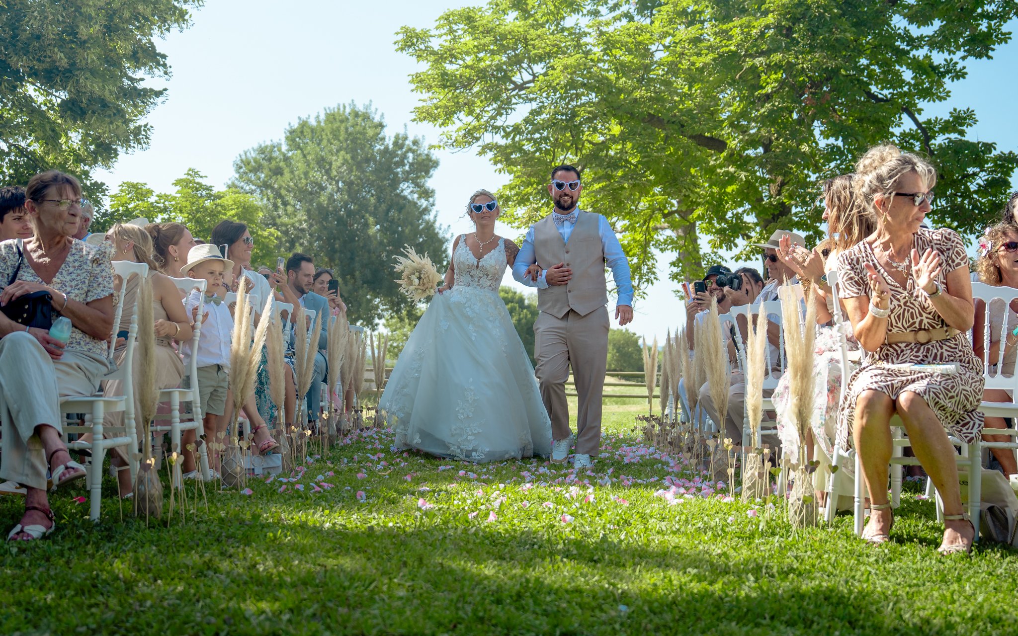 Un couple de mariés marchant dans un allée lors d'une cérémonie de mariage à l'extérieur sous un grand arbre, entouré de invités assis et pris en photo.
