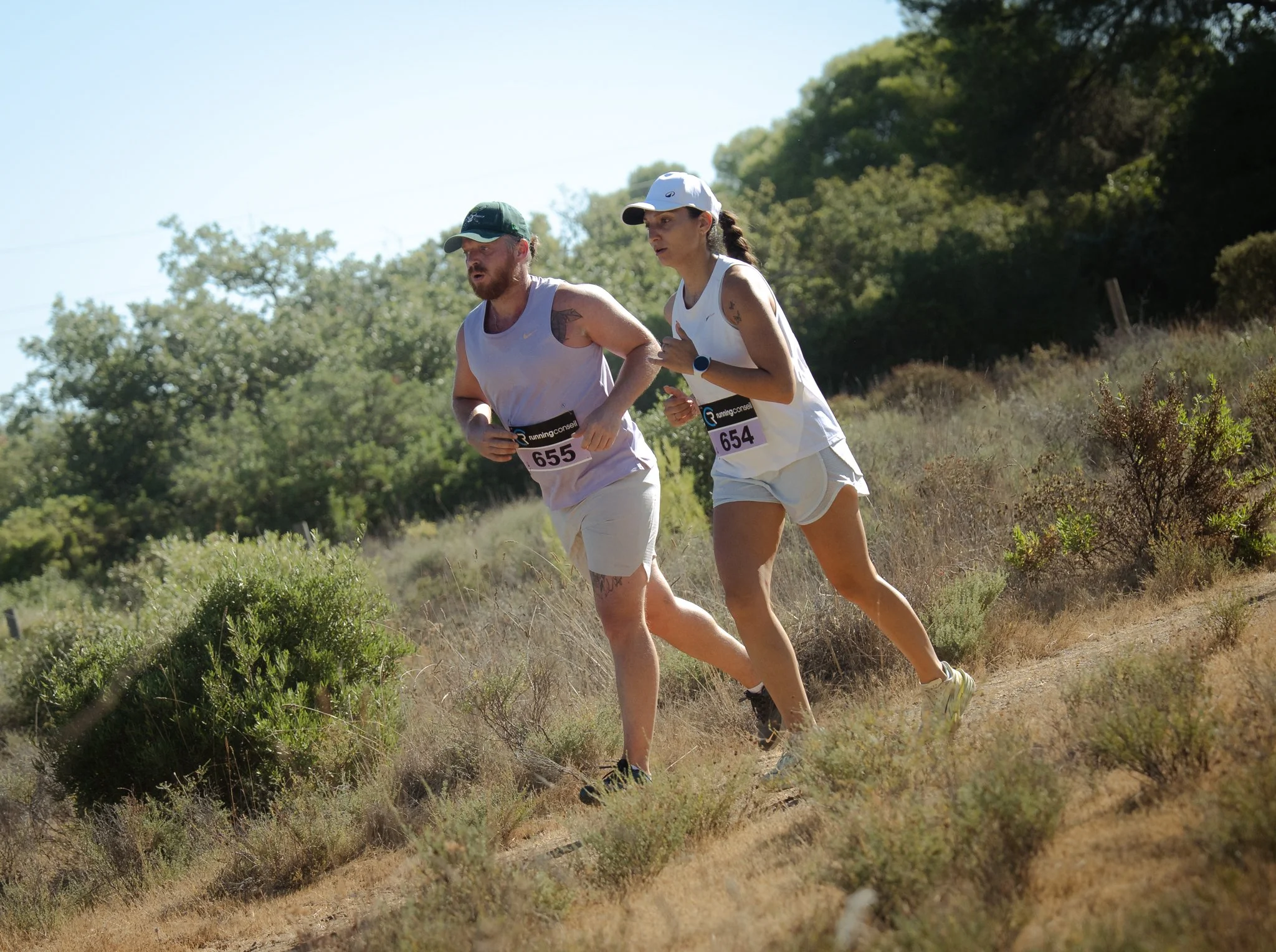 Deux coureurs, un homme et une femme, participent à une course en plein air dans un environnement naturel avec des arbres et des buissons, portant des vêtements de course et des numéros.