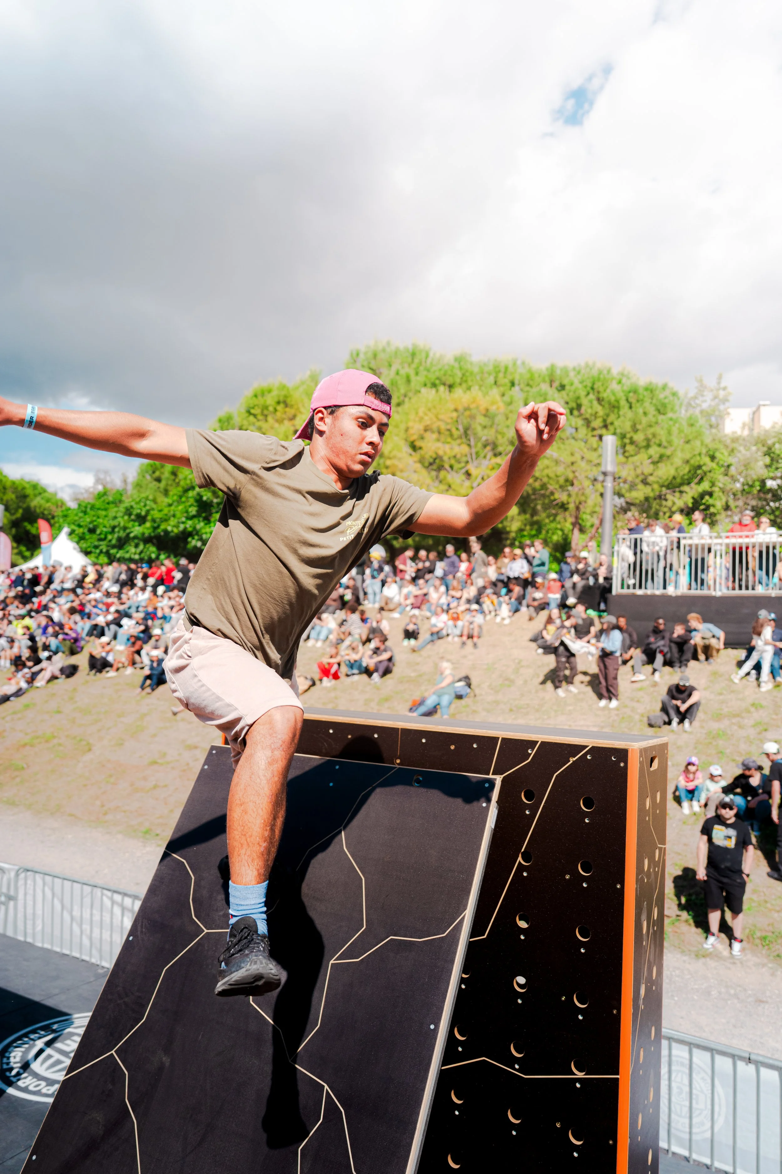 Un jeune homme portant une casquette rose et un t-shirt beige, en train d'escalader une structure d'escalade en bois avec des trous, lors d'un événement en plein air, avec un grand nombre de spectateurs et un ciel nuageux en arrière-plan.