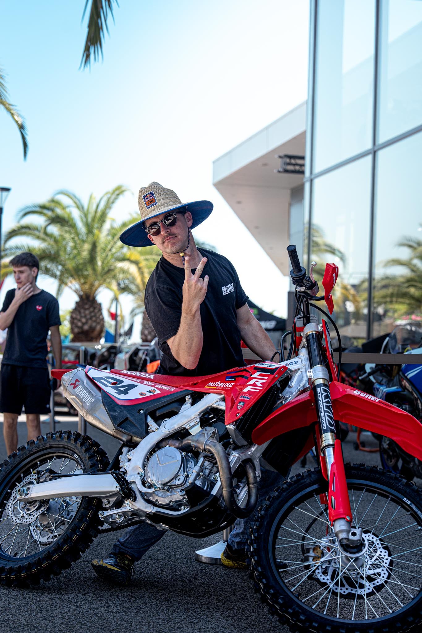 A man in a wide-brimmed hat and sunglasses standing next to a red off-road motorcycle, making a peace sign with his finger, with palm trees and a modern building in the background.