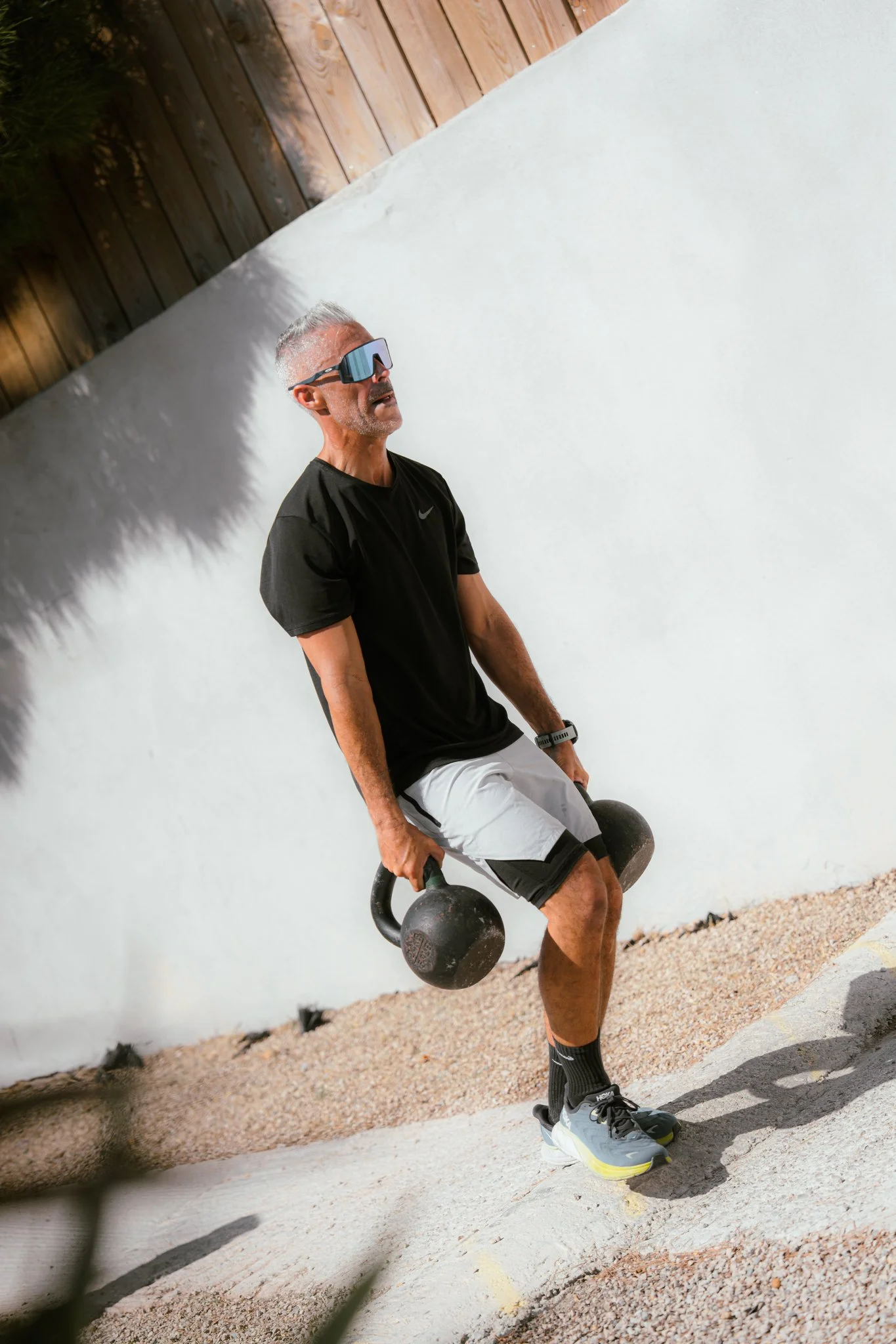Homme pèse avec kettlebells, portant lunettes de sport, chemise noire et shorts, en extérieur contre un mur blanc en bois.