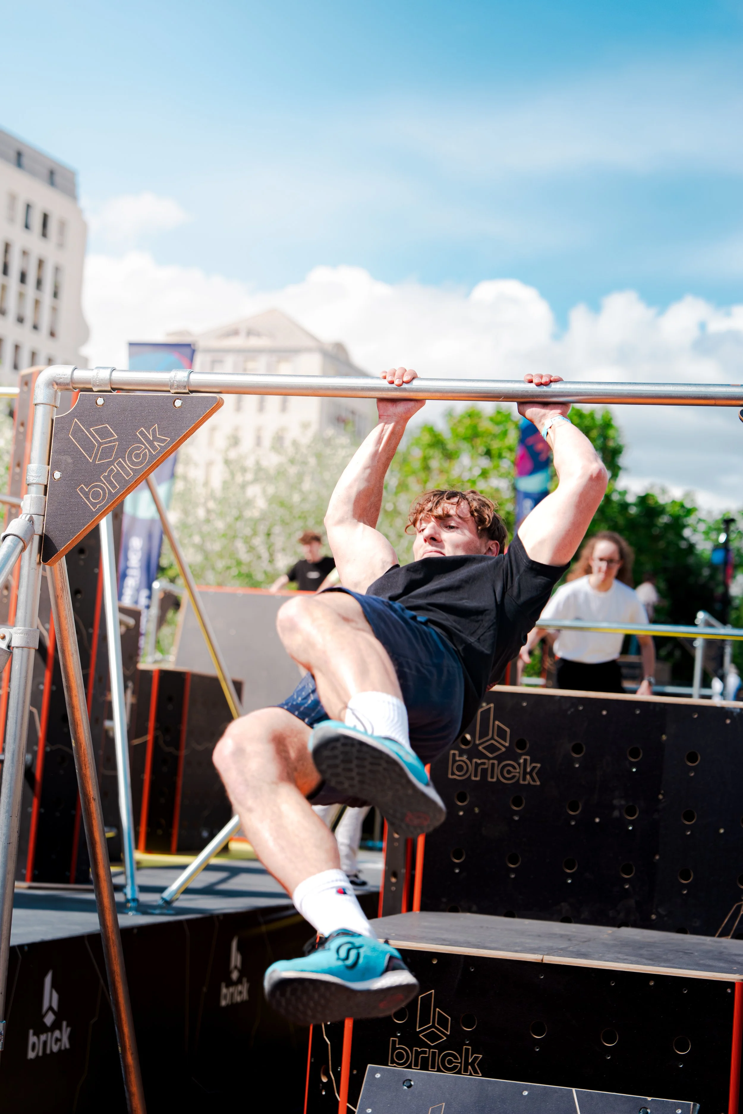 Un homme pratique les exercices de pull-up lors d'un événement en plein air, avec un ciel bleu et des bâtiments en arrière-plan.