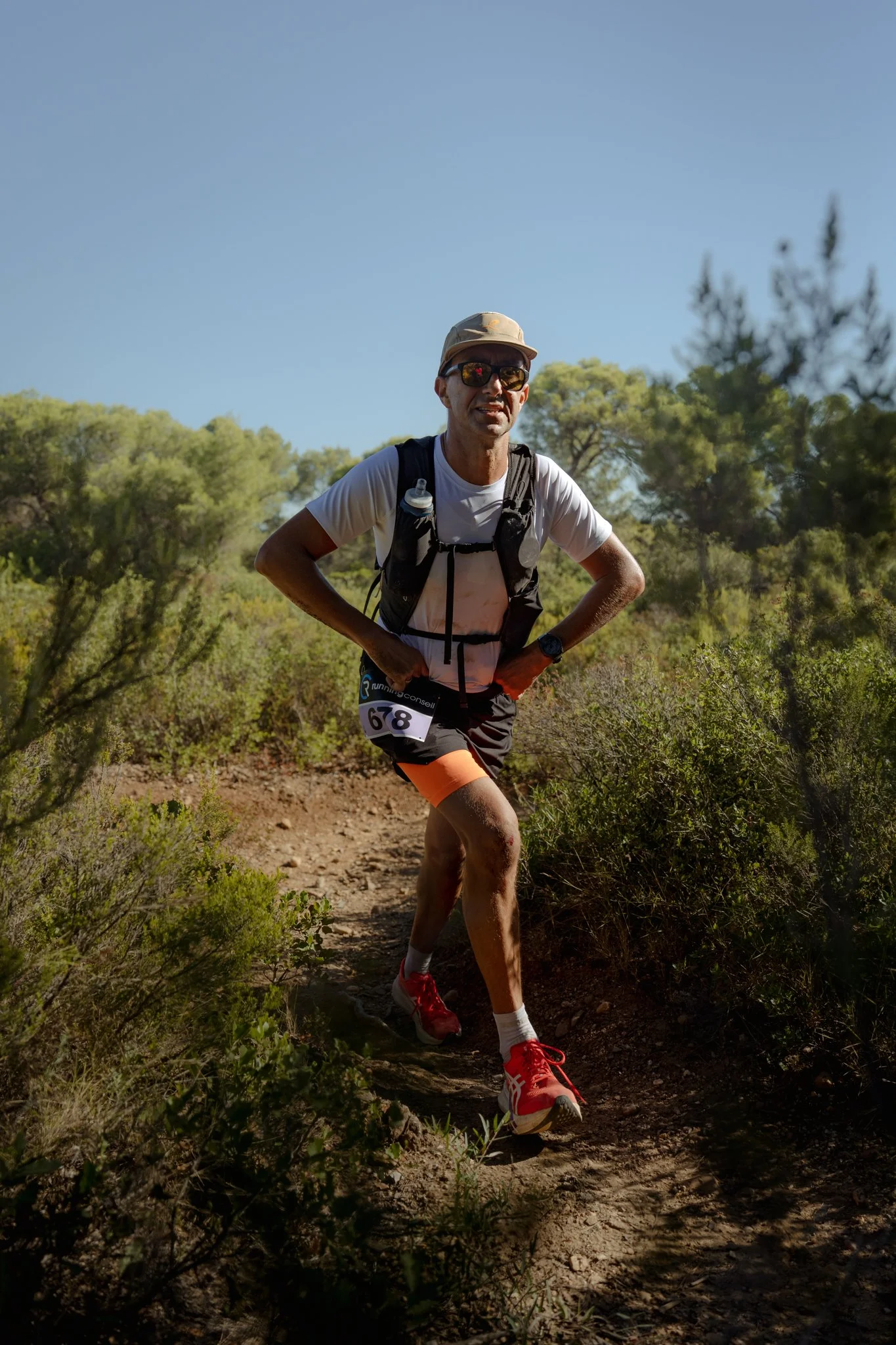 Un homme courant sur un sentier en pleine nature, portant un sac à dos, des lunettes de soleil et une casquette, avec un numéro de course 678, entouré d'arbres et de végétation sous un ciel clair.