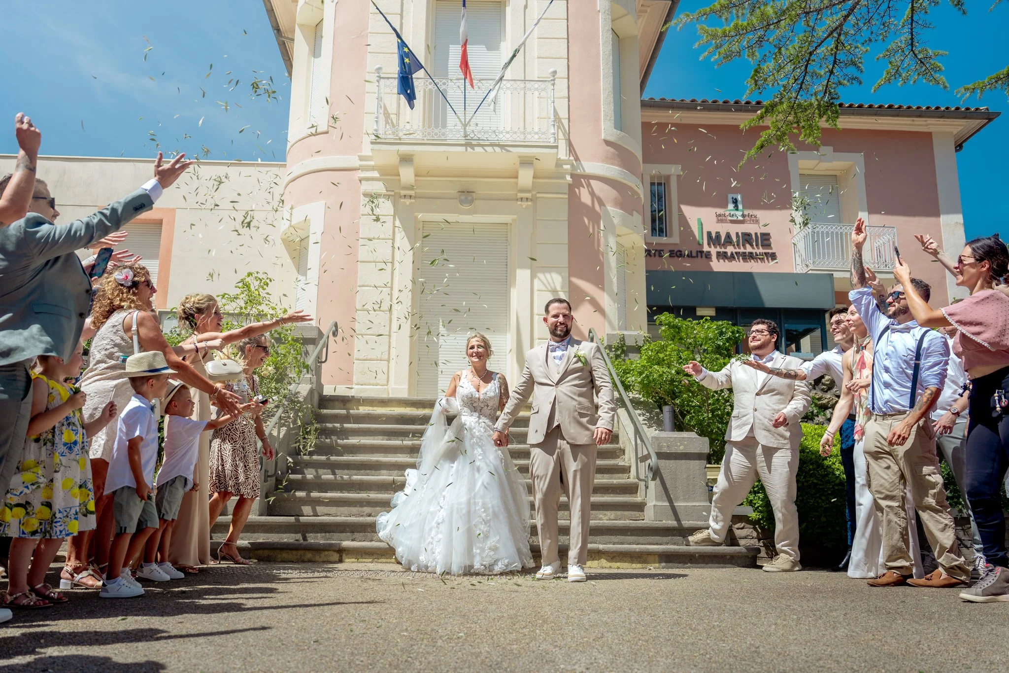 Un couple marié descend les escaliers de l'hôtel de ville avec des invités autour, en pleine célébration de mariage par une belle journée ensoleillée.