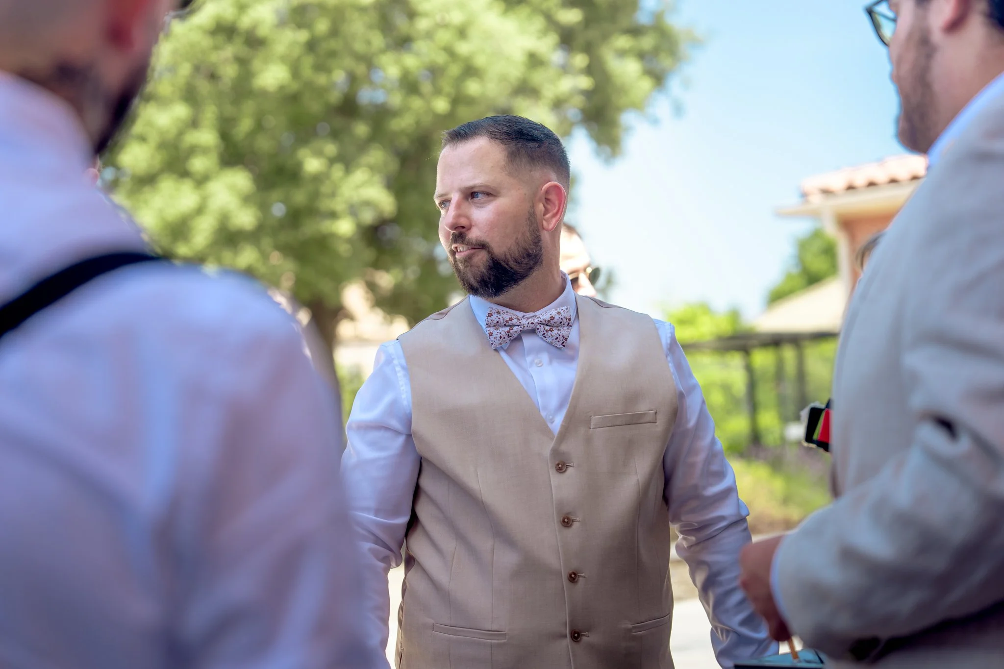 Un homme avec une barbe, portant un gilet beige et un noeud papillon à motifs, parle à deux autres personnes lors d'une réunion en extérieur avec des arbres verts et un ciel bleu en arrière-plan.
