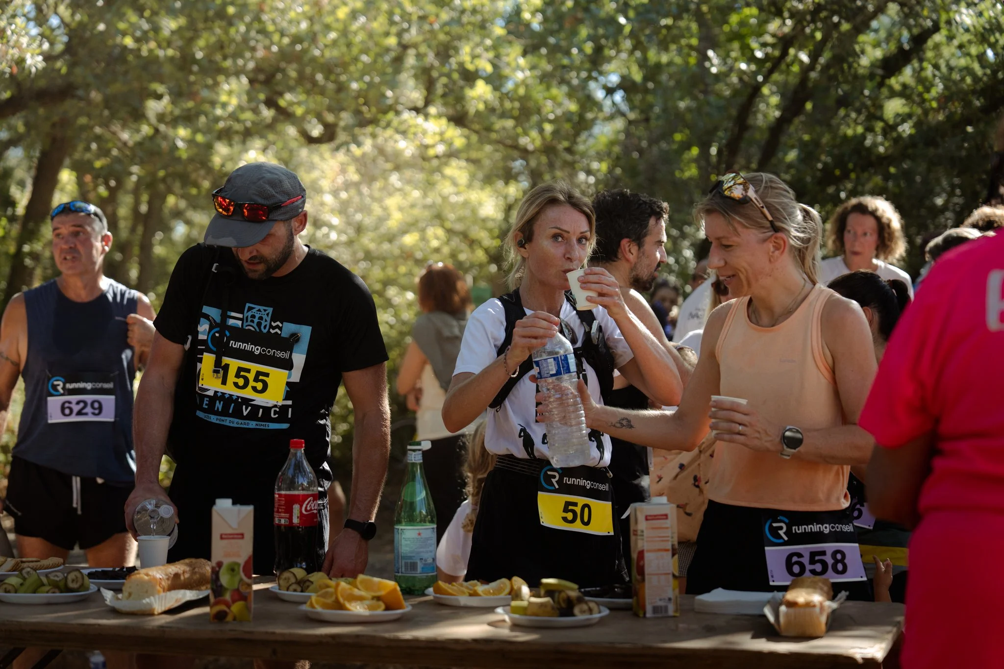 Groupe de personnes participant à une course ou une course caritative, distribuant et recevant des refreshments à l'arrivée, avec des tables remplies de nourriture et de boissons, dans un environnement en plein air avec des arbres en arrière-plan.