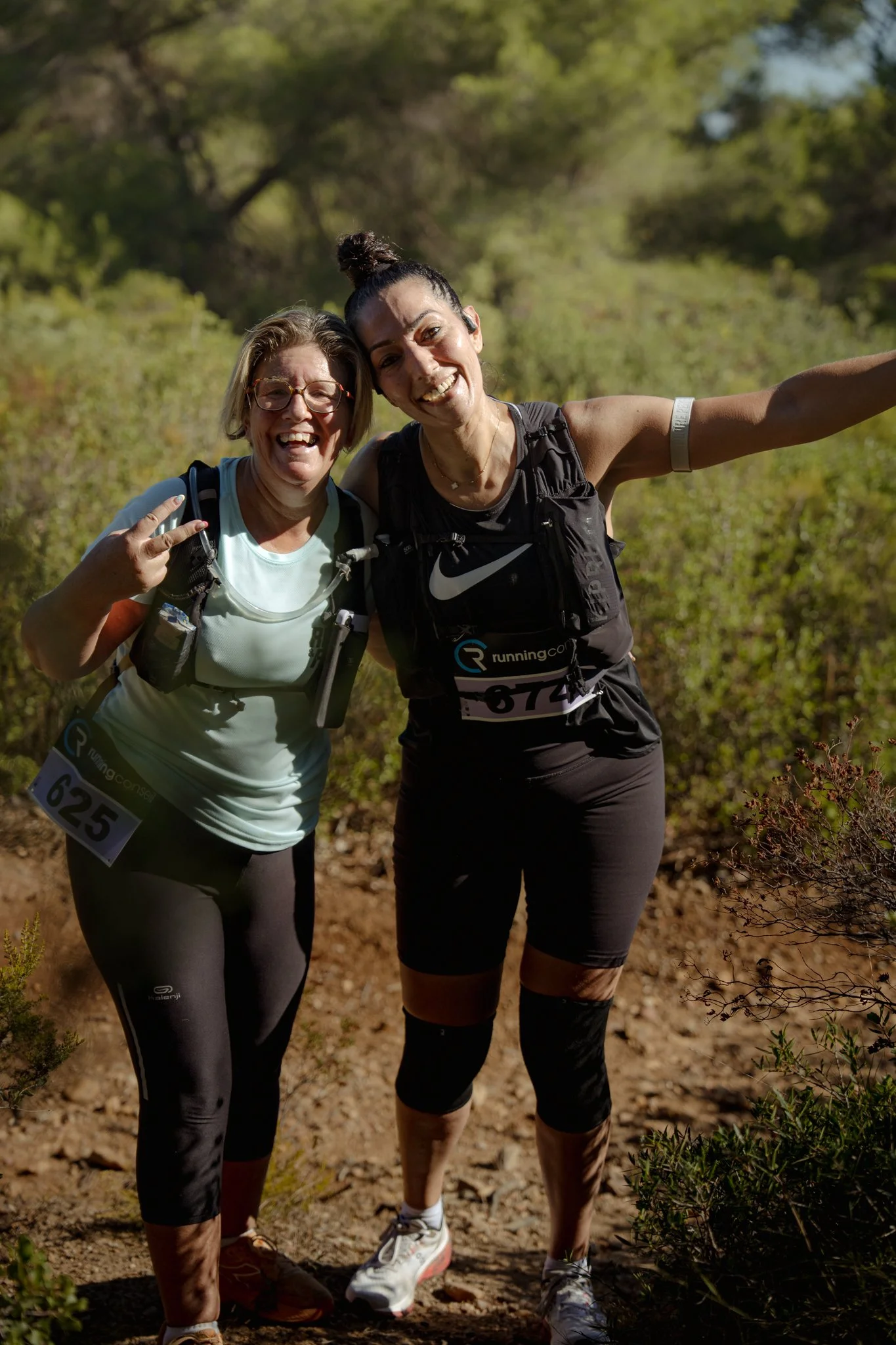 Deux femmes souriantes en vêtements de course posent dans un forêt naturelle, avec des numéros de course attachés à leur vêtements.