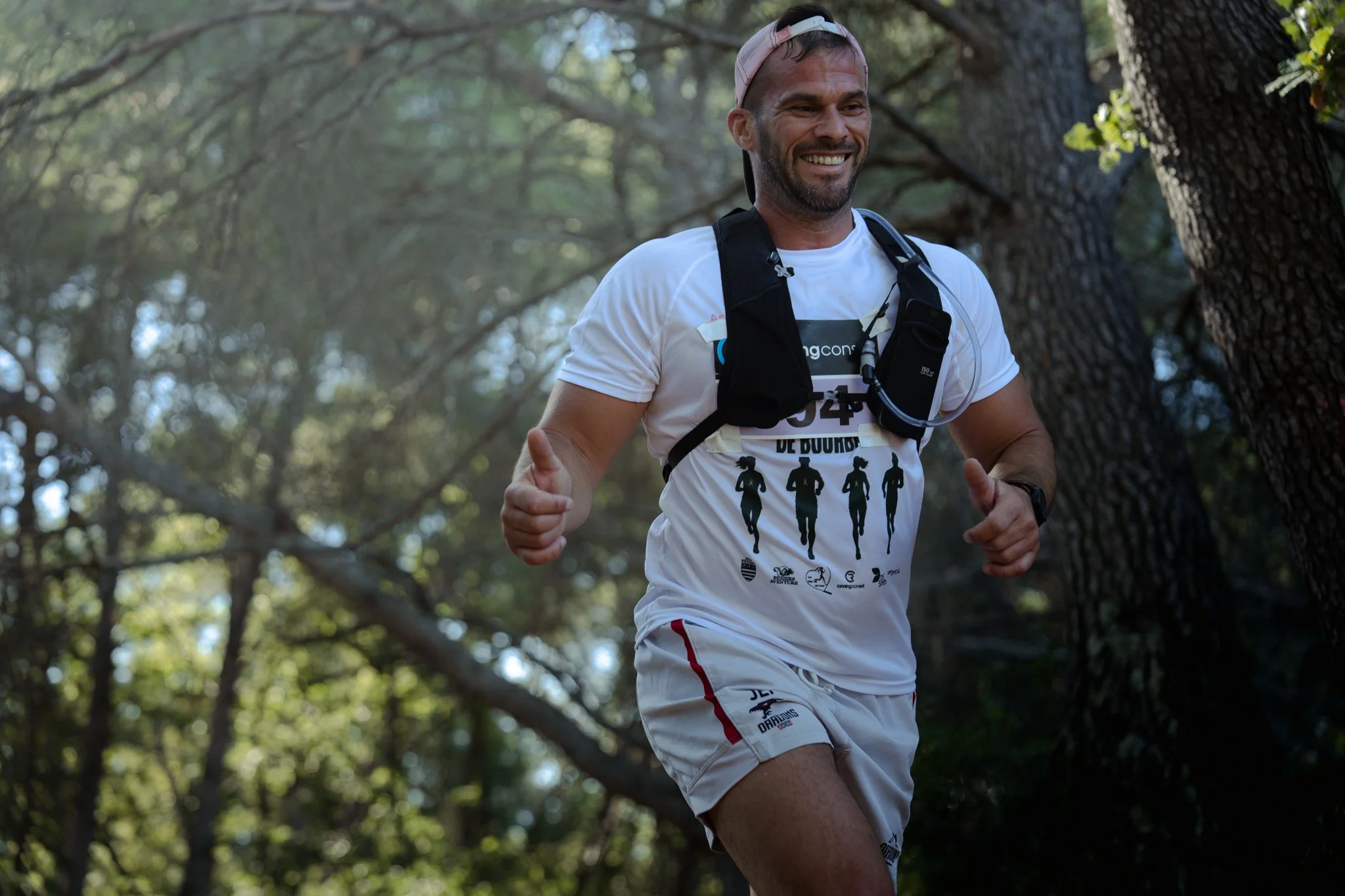 Homme souriant courant dans une forêt, portant un t-shirt blanc avec des silhouettes de runners, une veste d'hydratation et une casquette