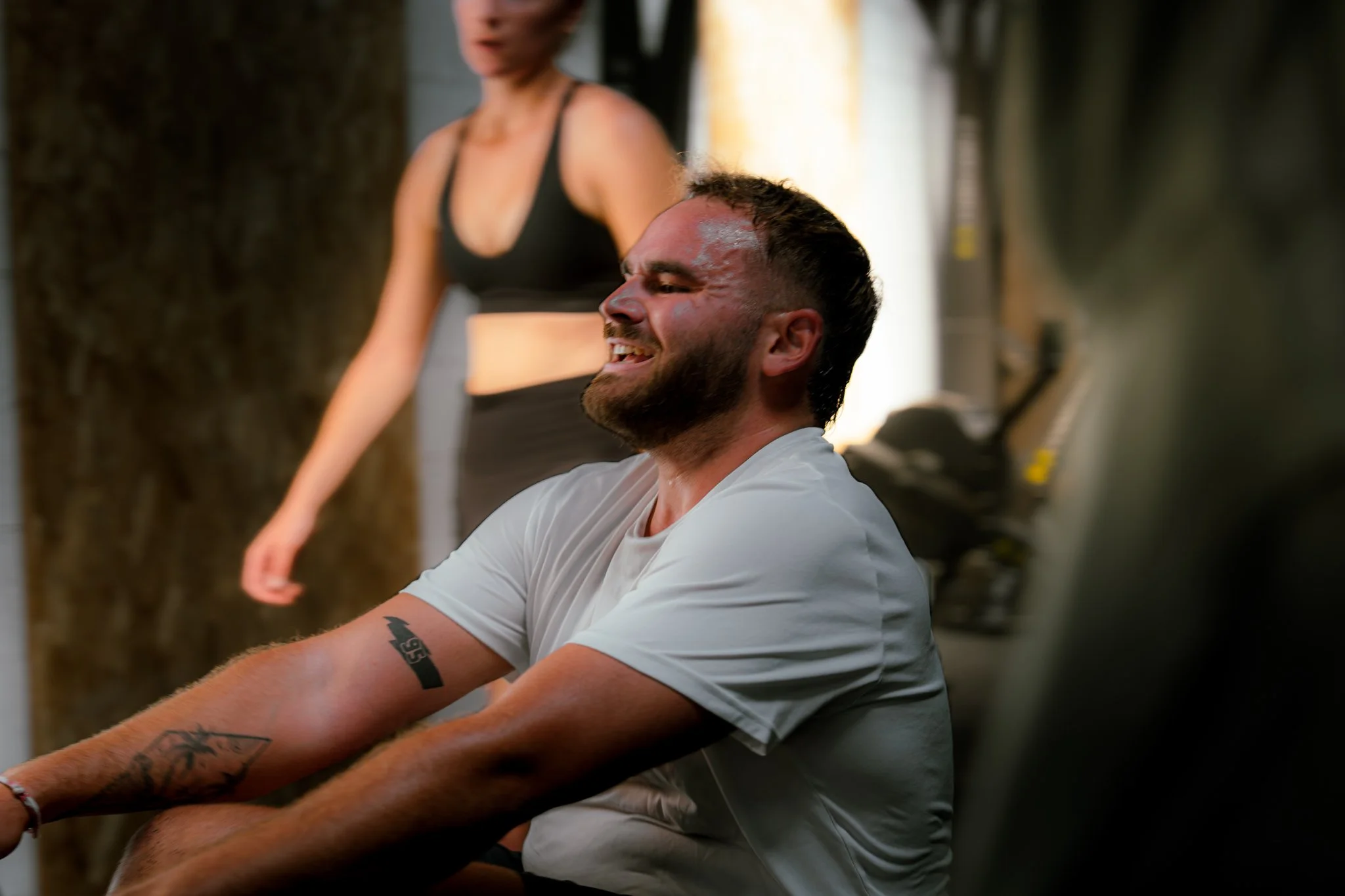 Un homme souriant, assis avec un bras tendu, dans un environnement intérieur, avec une femme en arrière-plan, floue, portant un haut noir.