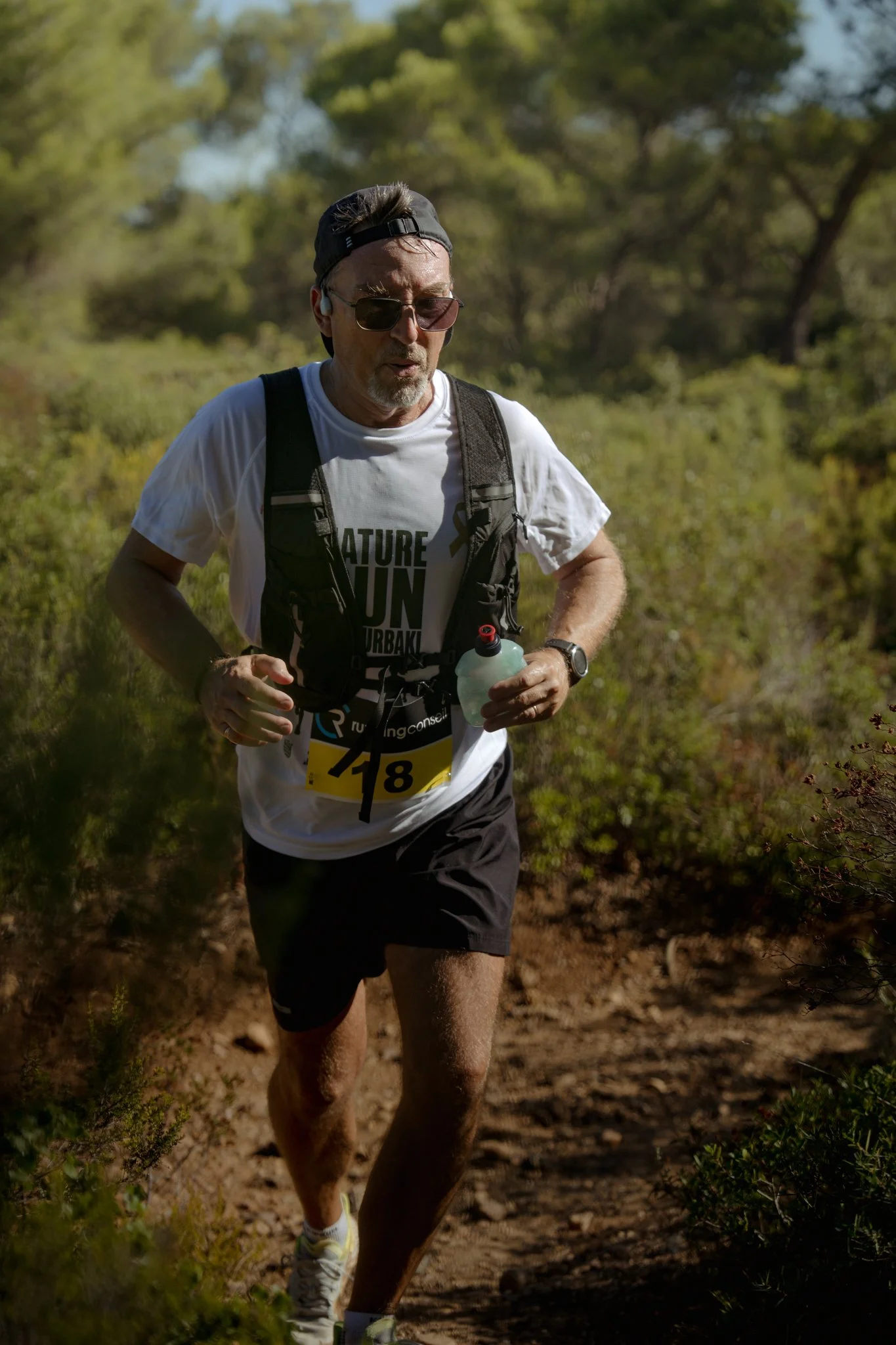 Un homme court sur un sentier de forêt en tenue de running, portant un t-shirt blanc, un sac à dos, des lunettes de soleil et une bouteille d'eau. Il semble concentré et déterminé.