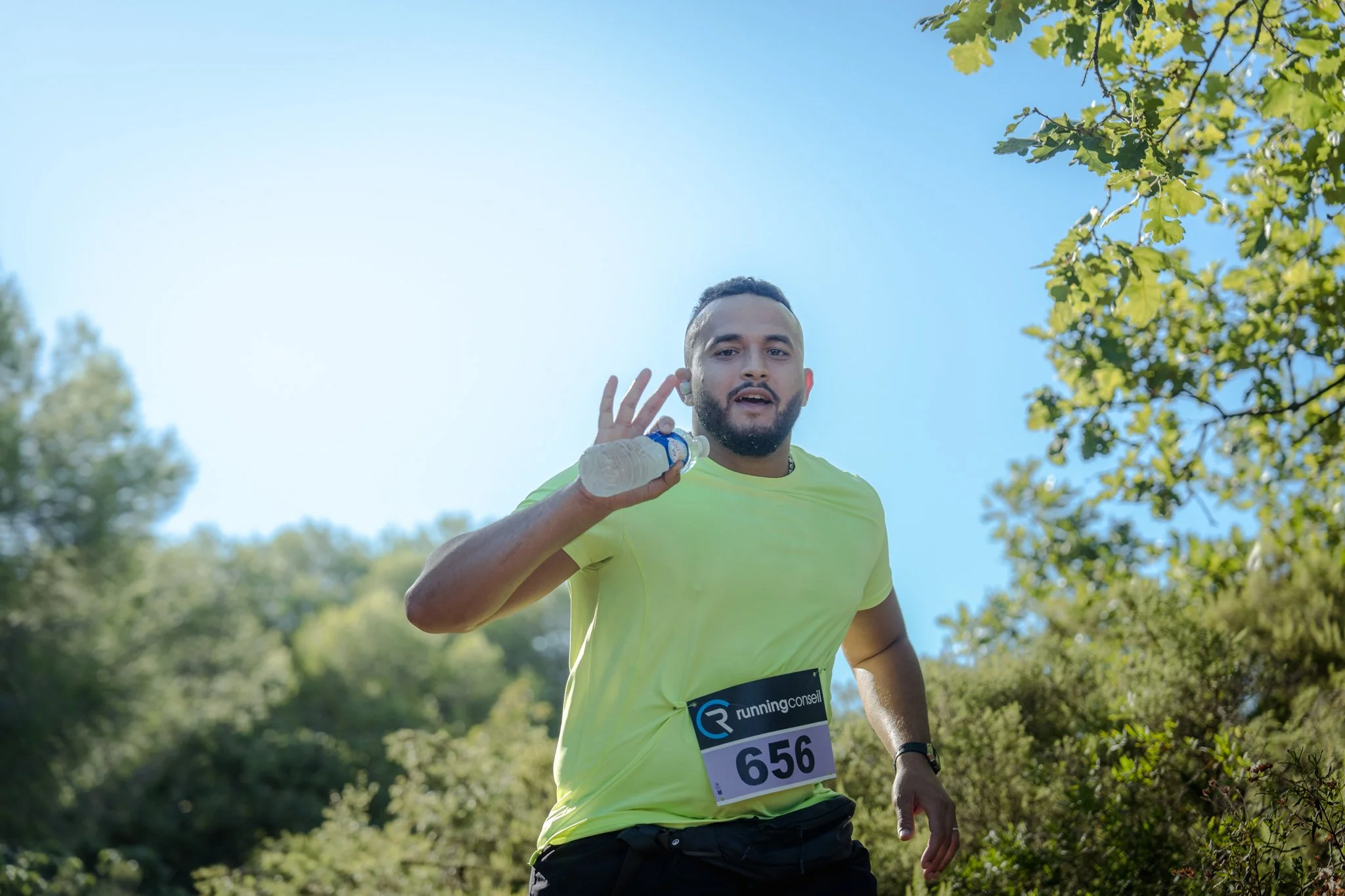 Homme courant en extérieur avec un bidon d'eau à la main, portant un t-shirt fluorescent et un numéro de course 656, sous un ciel bleu avec des arbres verts.