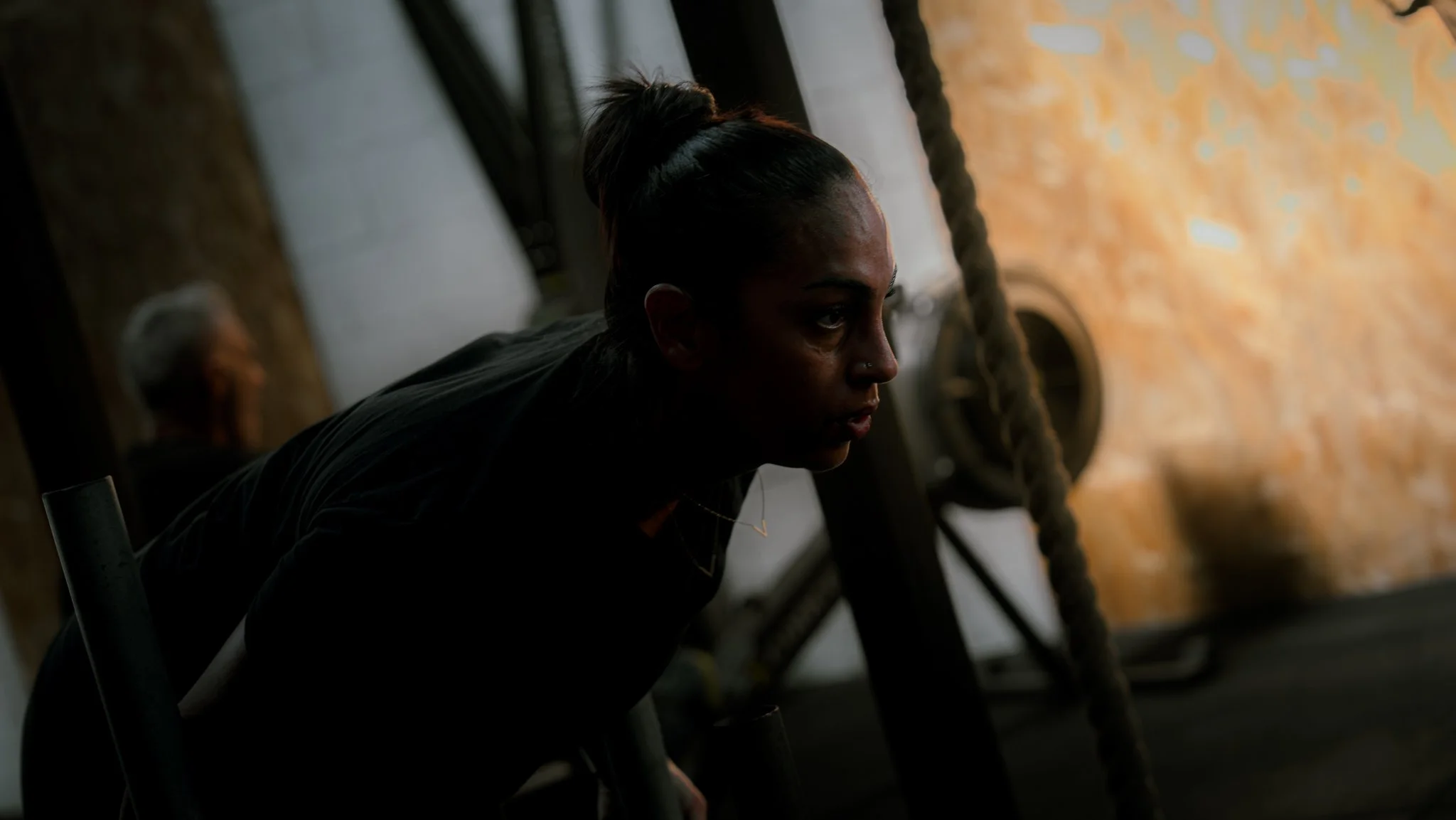 Une femme aux cheveux attachés en chignon, portant un vêtements noirs, semble concentrée, dans une salle de musculation ou à un entraînement.