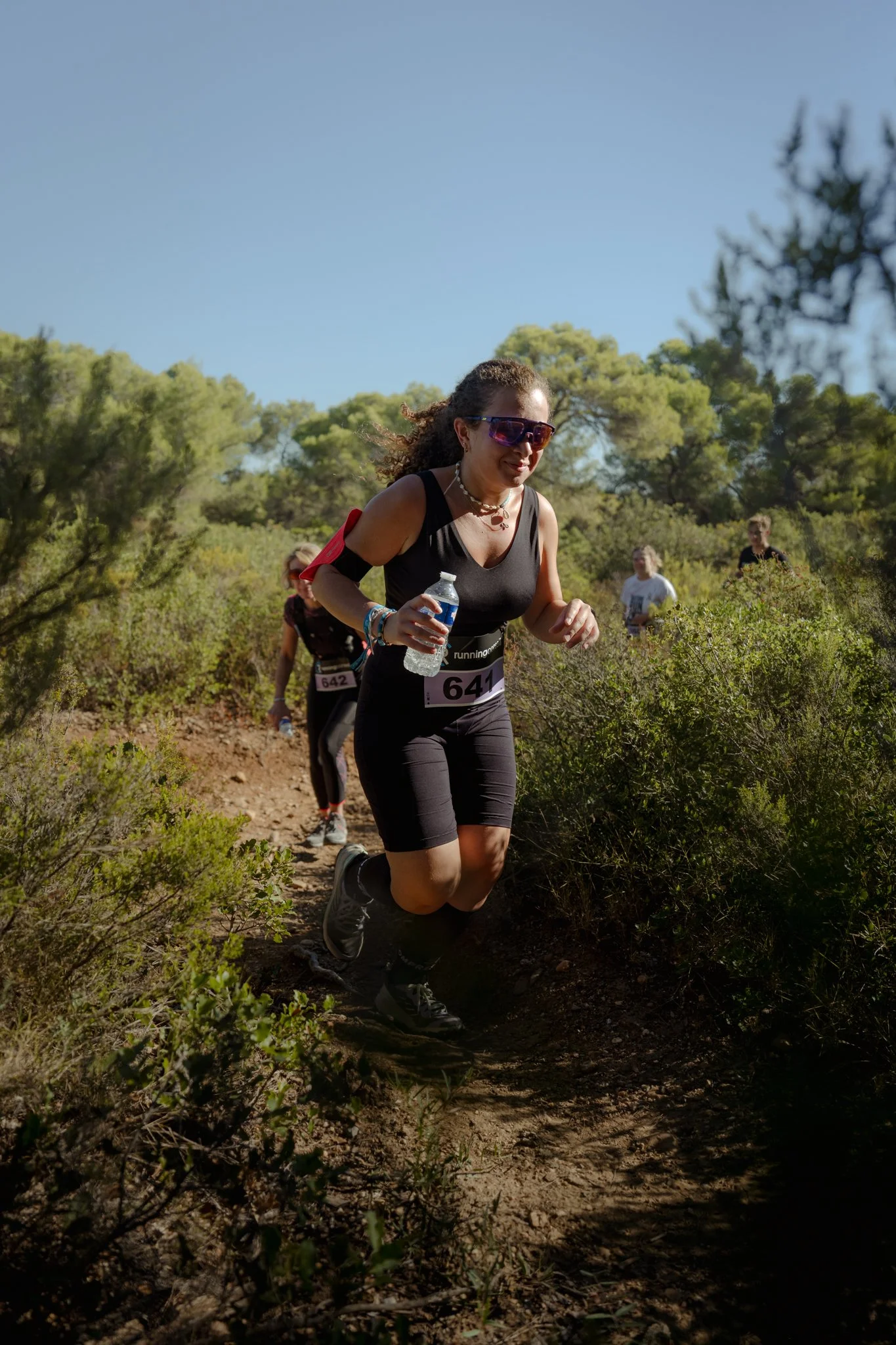 Femme courant dans une forêt, portant un numéro de course 641, lunettes de soleil, bouteille d'eau, cheveux bouclés, en robe noire, sous un ciel bleu.