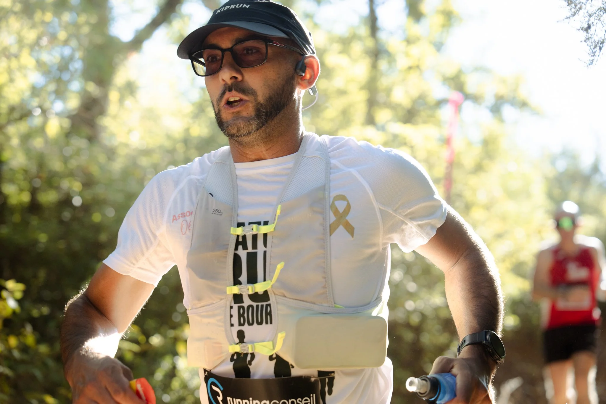 Un homme portant une casquette, des lunettes, un t-shirt blanc et une veste de course participe à une course en plein air, avec des arbres en arrière-plan.