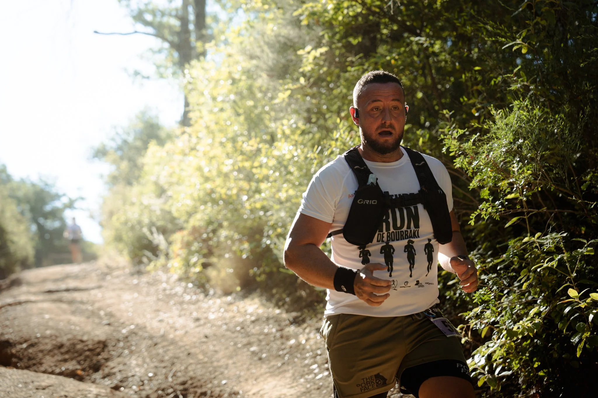 Un homme court dans un sentier forestier ensoleillé, portant une tenue de running et une bouteille d'eau. Il porte un t-shirt blanc avec une illustration de plusieurs coureurs et un sac à dos. Background de feuillage vert et des arbres.