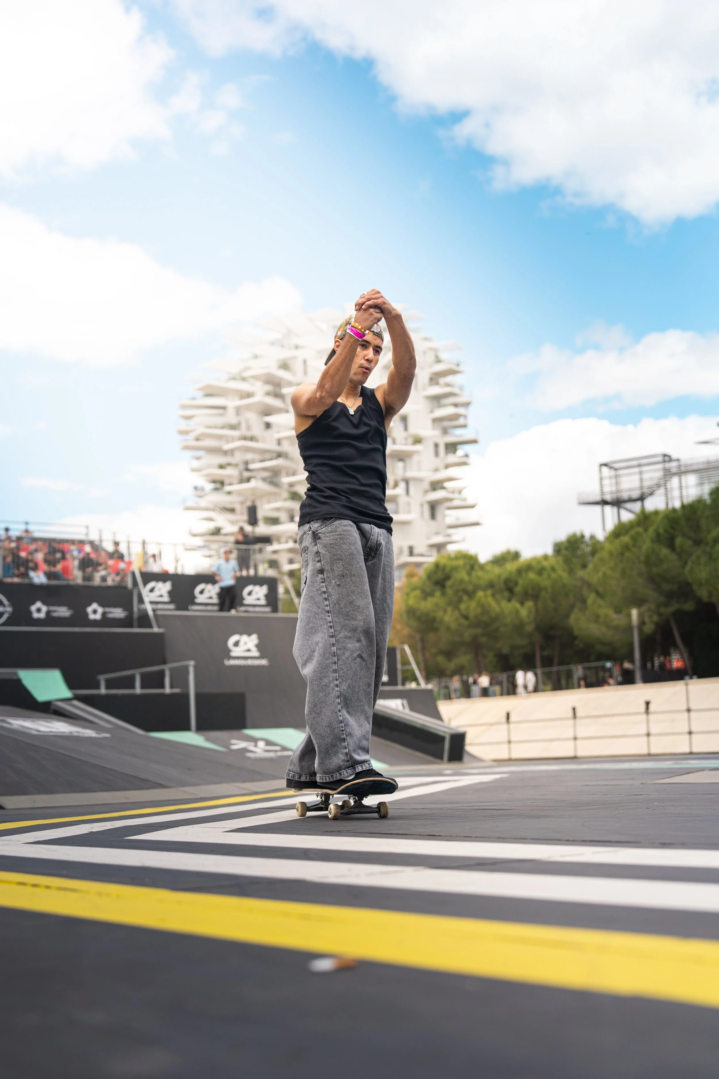 Jeune homme qui fait du skateboard dans un skatepark en plein air avec un bâtiment moderne en arrière-plan et des spectateurs