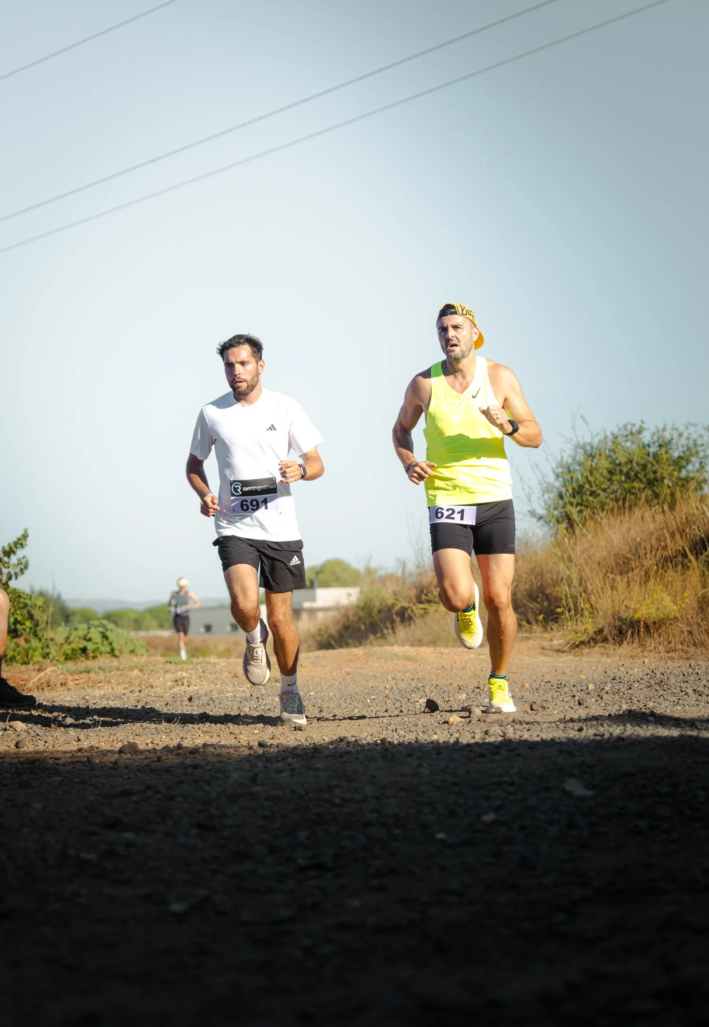 Deux hommes courent sur un sentier en plein air, portant des vêtements de sport avec des numéros de course, sous un ciel dégagé.