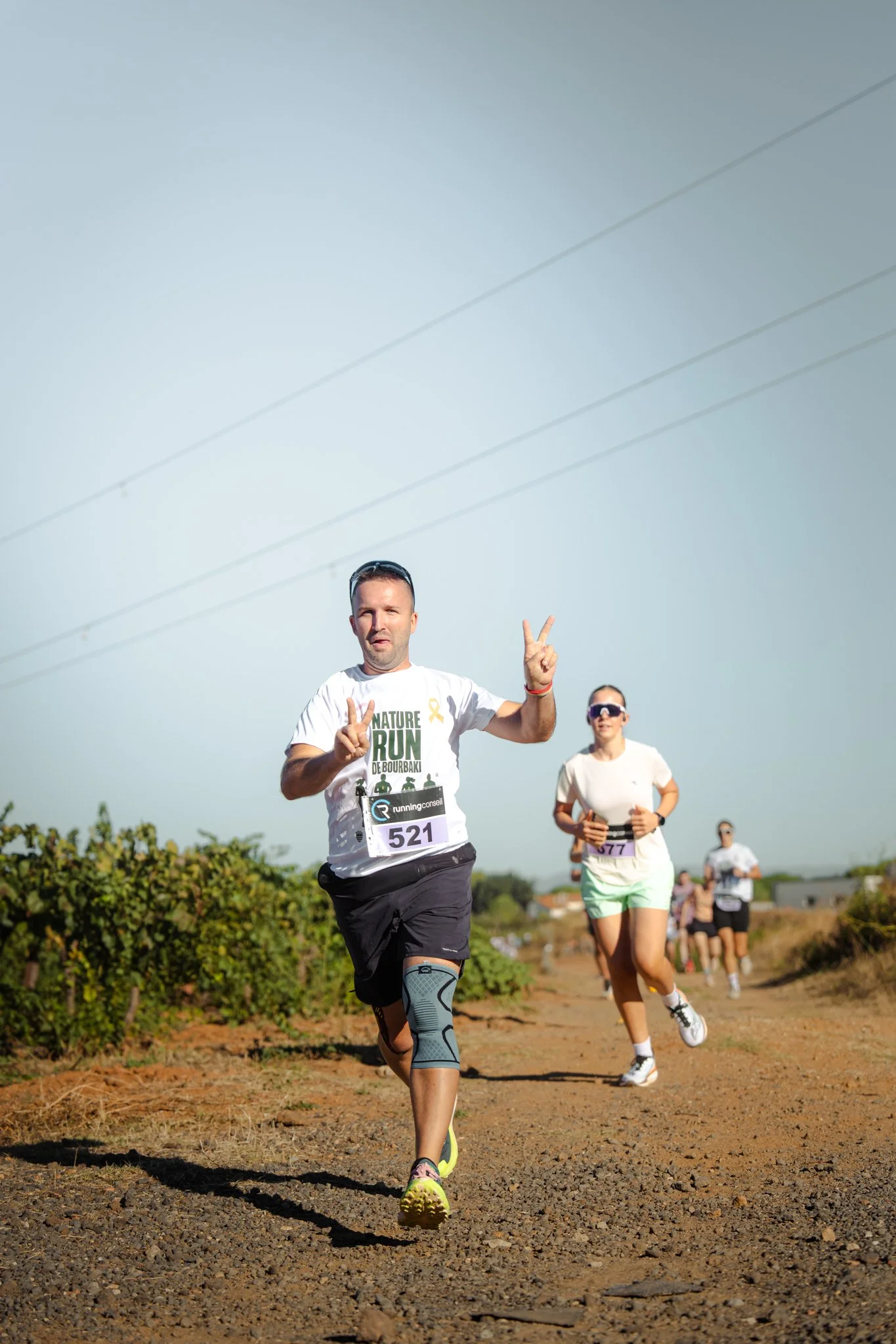 Homme courant lors d'un marathon ou course en plein air, portant un t-shirt blanc avec inscriptions, numéro 521, et une jambe prothétique, avec une femme et d'autres coureurs derrière, dans un paysage rural ensoleillé.