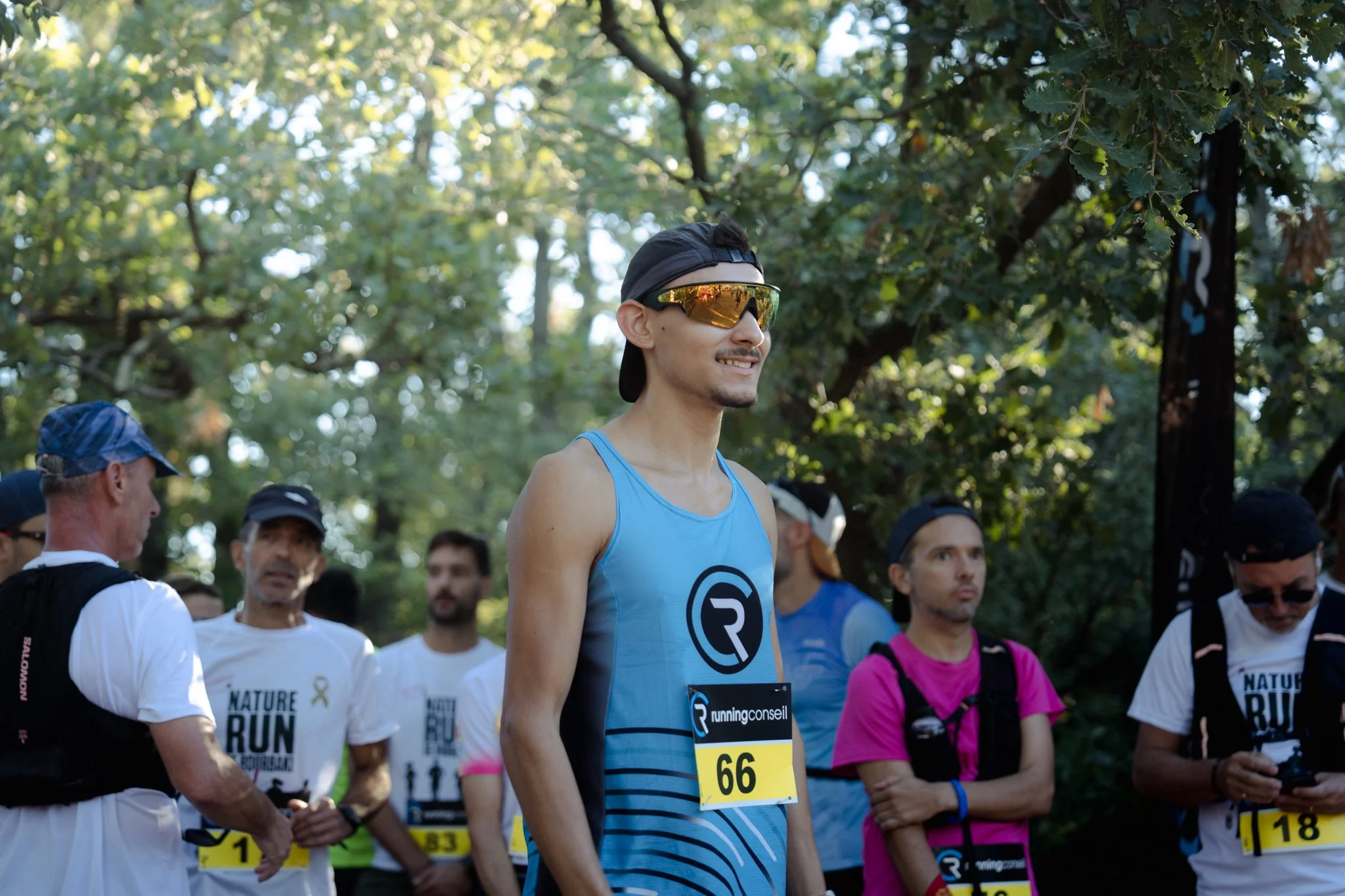Homme portant des lunettes de soleil réfléchissantes, un débardeur bleu avec un logo, participant à une course dans un parc avec d'autres coureurs autour.