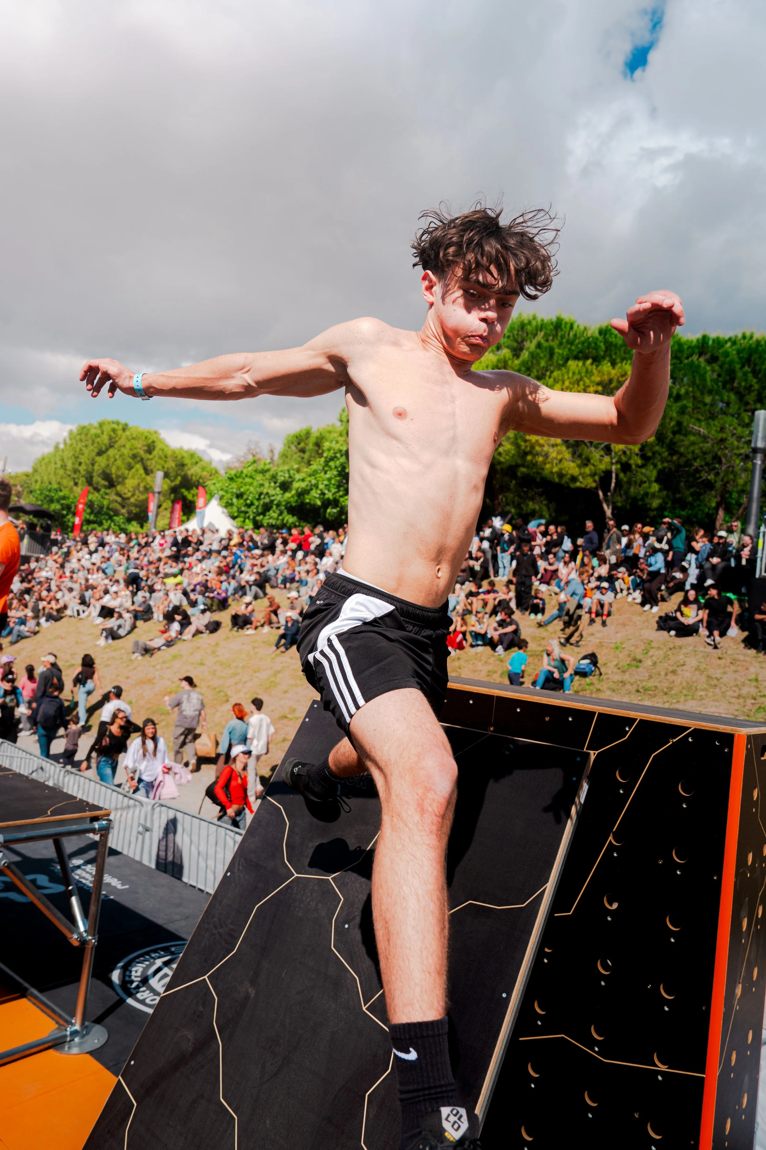Un jeune homme torse nu avec une expression concentrée, en train de faire une activité de saut ou d'exercice sur une plateforme en extérieur, avec un grand public dans le fond sous un ciel nuageux.