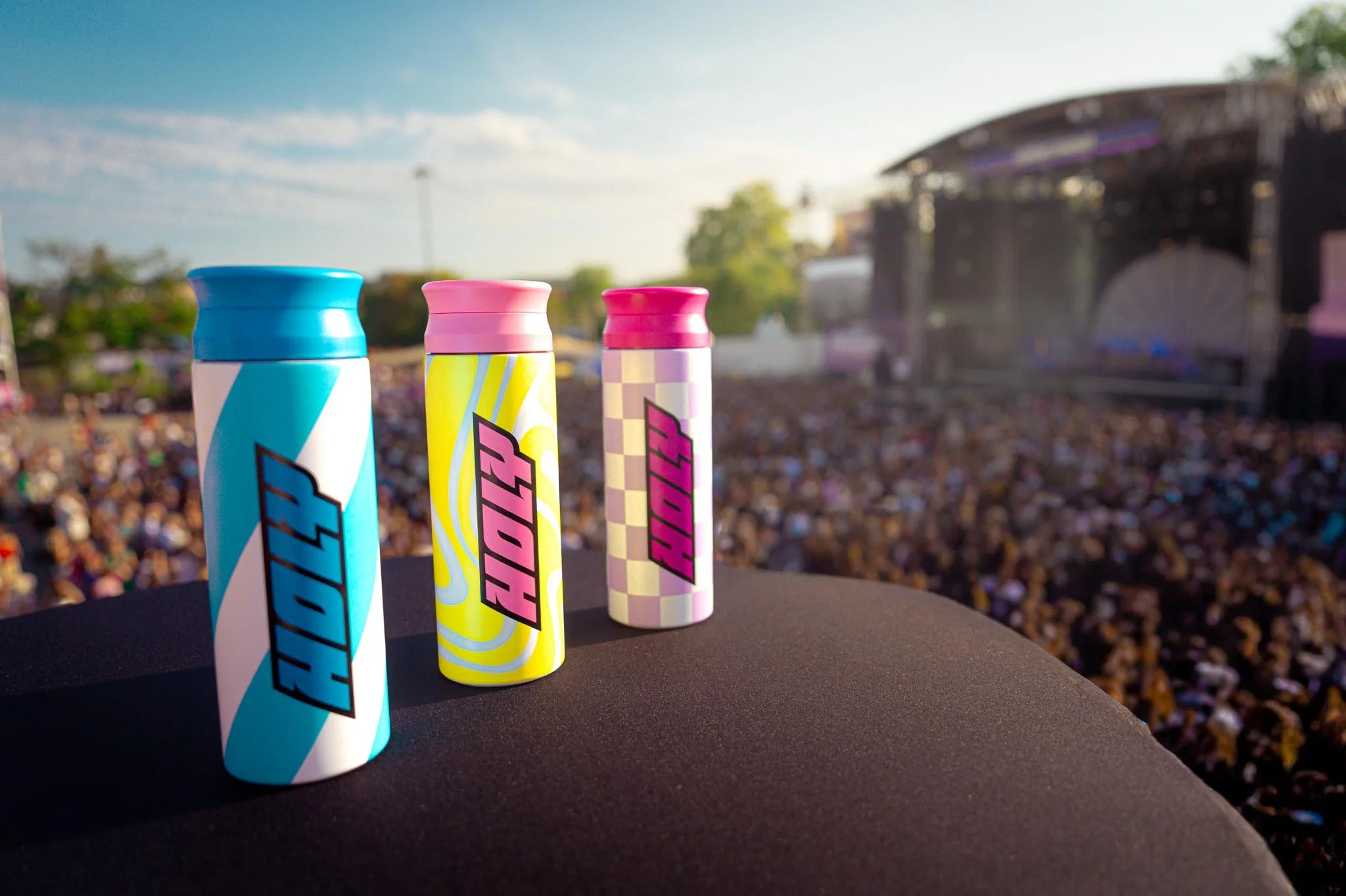 Three colorful cans with bold designs and text, standing on a black surface outdoors during daylight, with a blurred crowd and stage in the background.