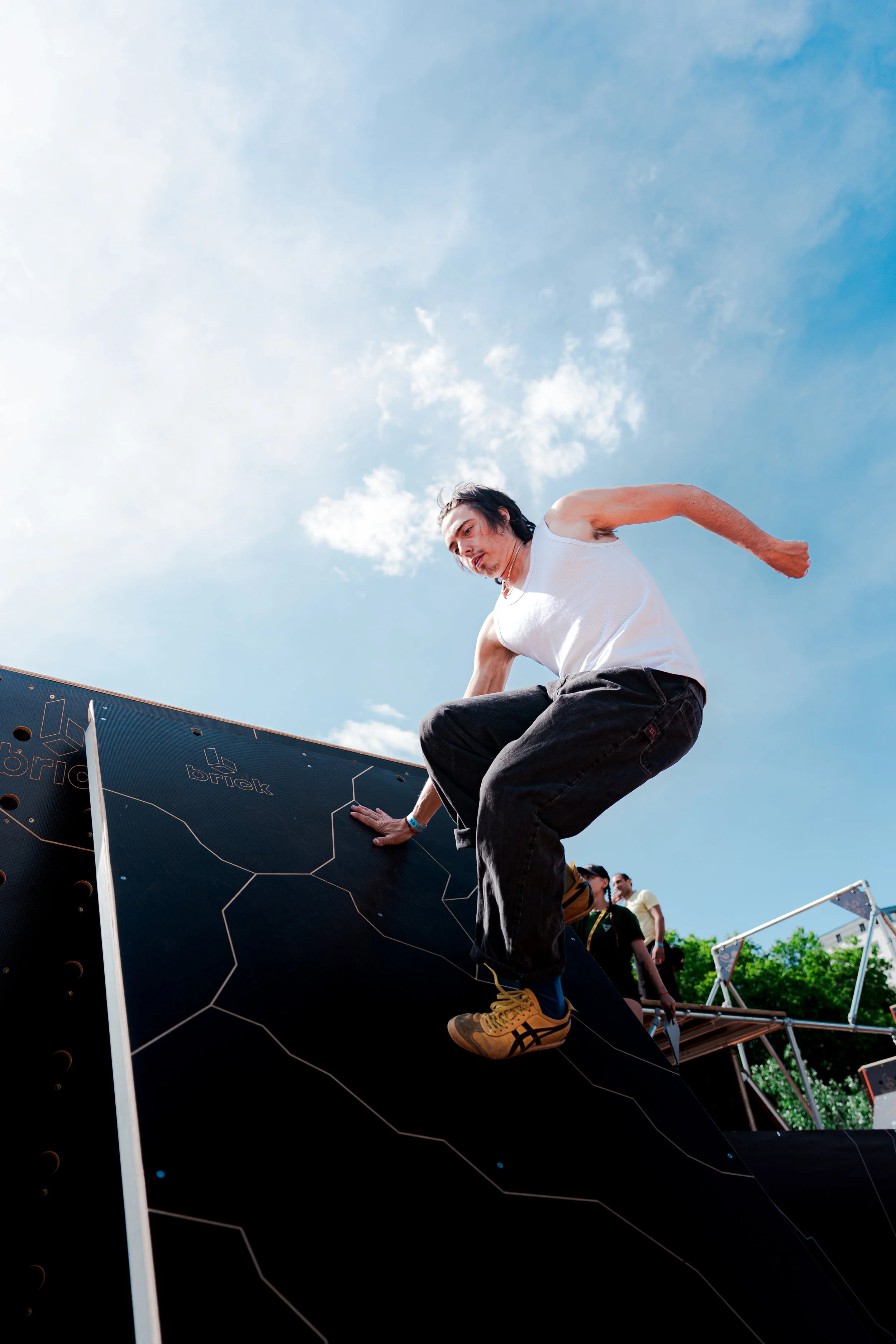Une personne grimpe sur une structure d'escalade en plein air, sous un ciel bleu avec quelques nuages, lors d'un événement ou d'un festival.
