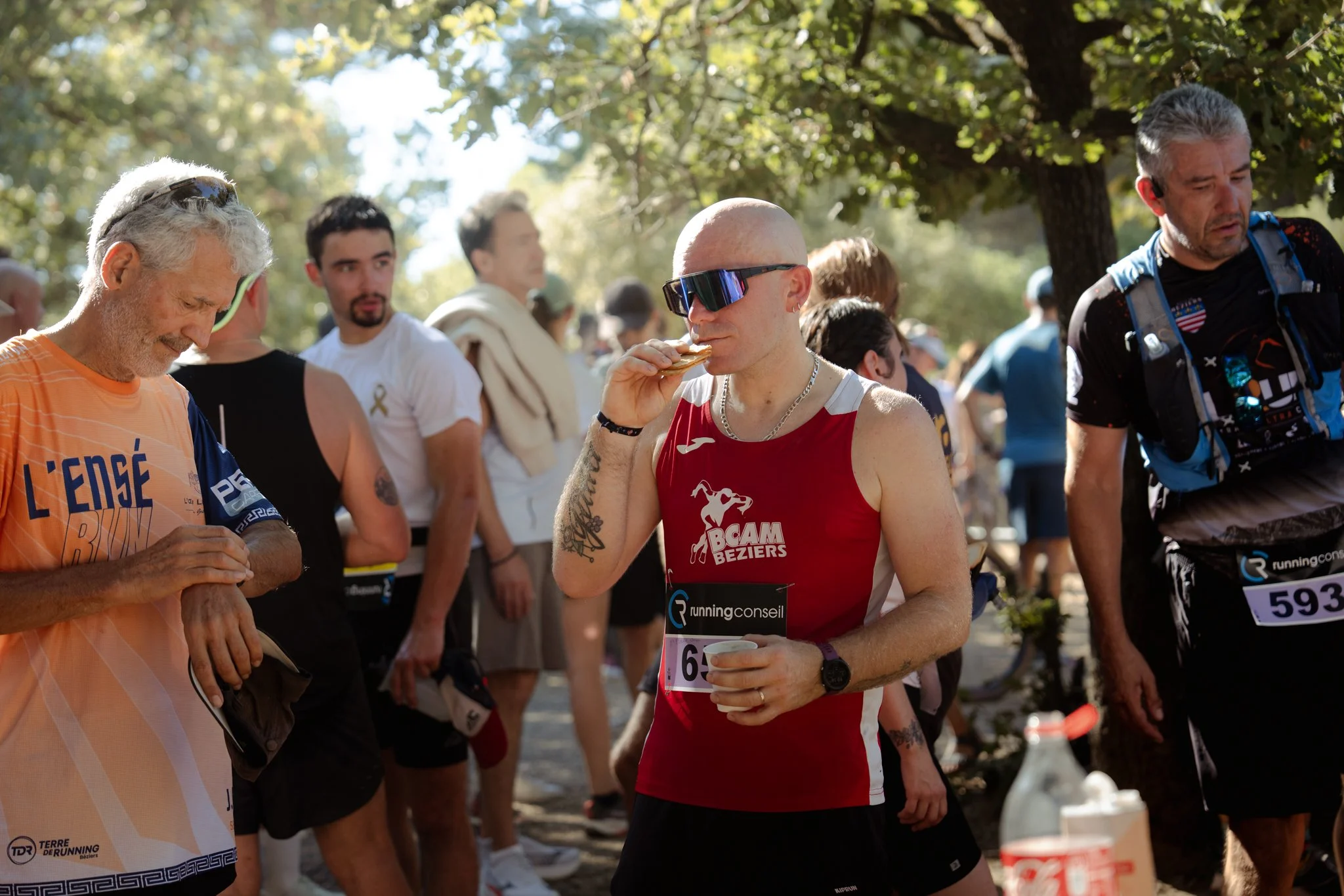 Des coureurs de course à pied se reposent et prennent une collation lors d'un événement en plein air, avec des arbres en arrière-plan.