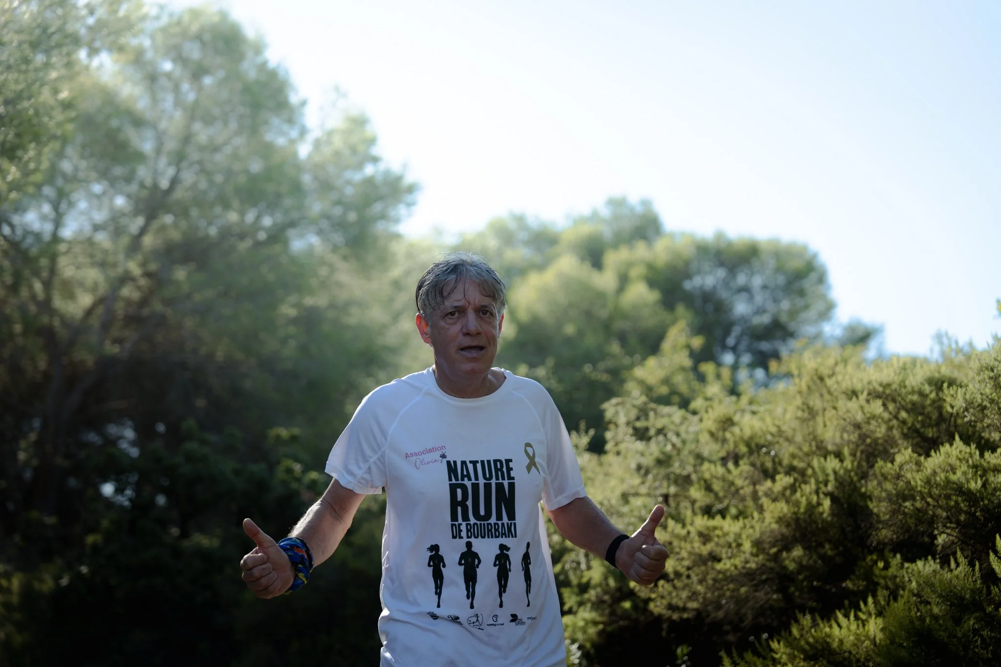 Homme courir en plein air, portant un t-shirt blanc à l'inscription 'NATURE RUN' et faisant un geste de victoire avec ses mains, dans un environnement verdoyant avec des arbres, sous un ciel ensoleillé.