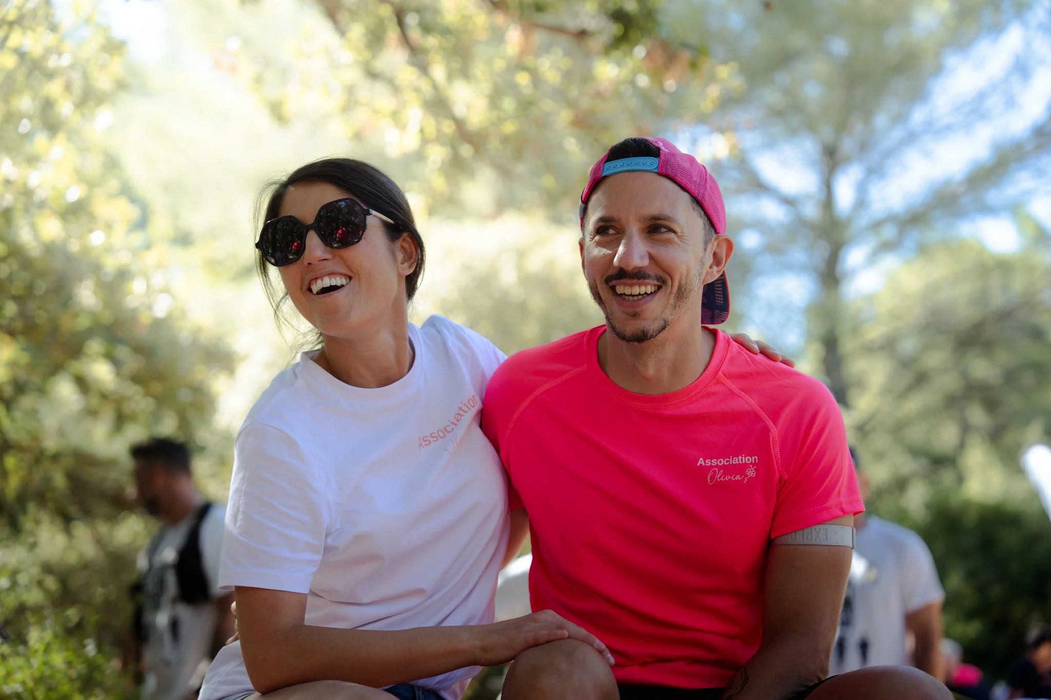 Deux personnes souriantes lors d'un événement en plein air, portant des t-shirts roses avec l'inscription 'Association Olivia', l'une porte des lunettes de soleil rondes et l'autre une casquette rose, entourées de verdure avec un ciel bleu.