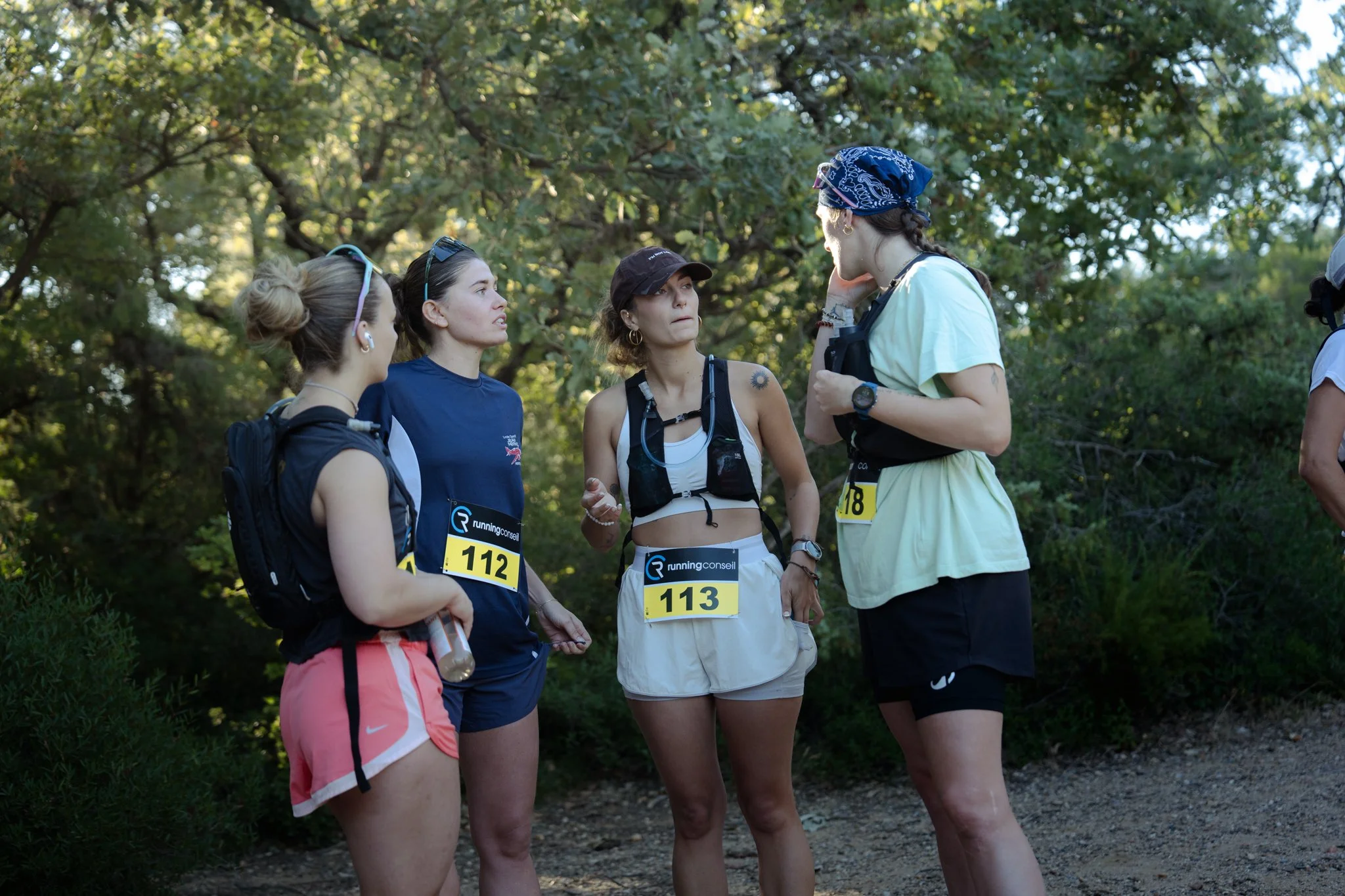 Un groupe de coureuses discute avec leur entraîneure dans un environnement boisée, portant des dossards, des vêtements de sport et des bouteilles d'eau.