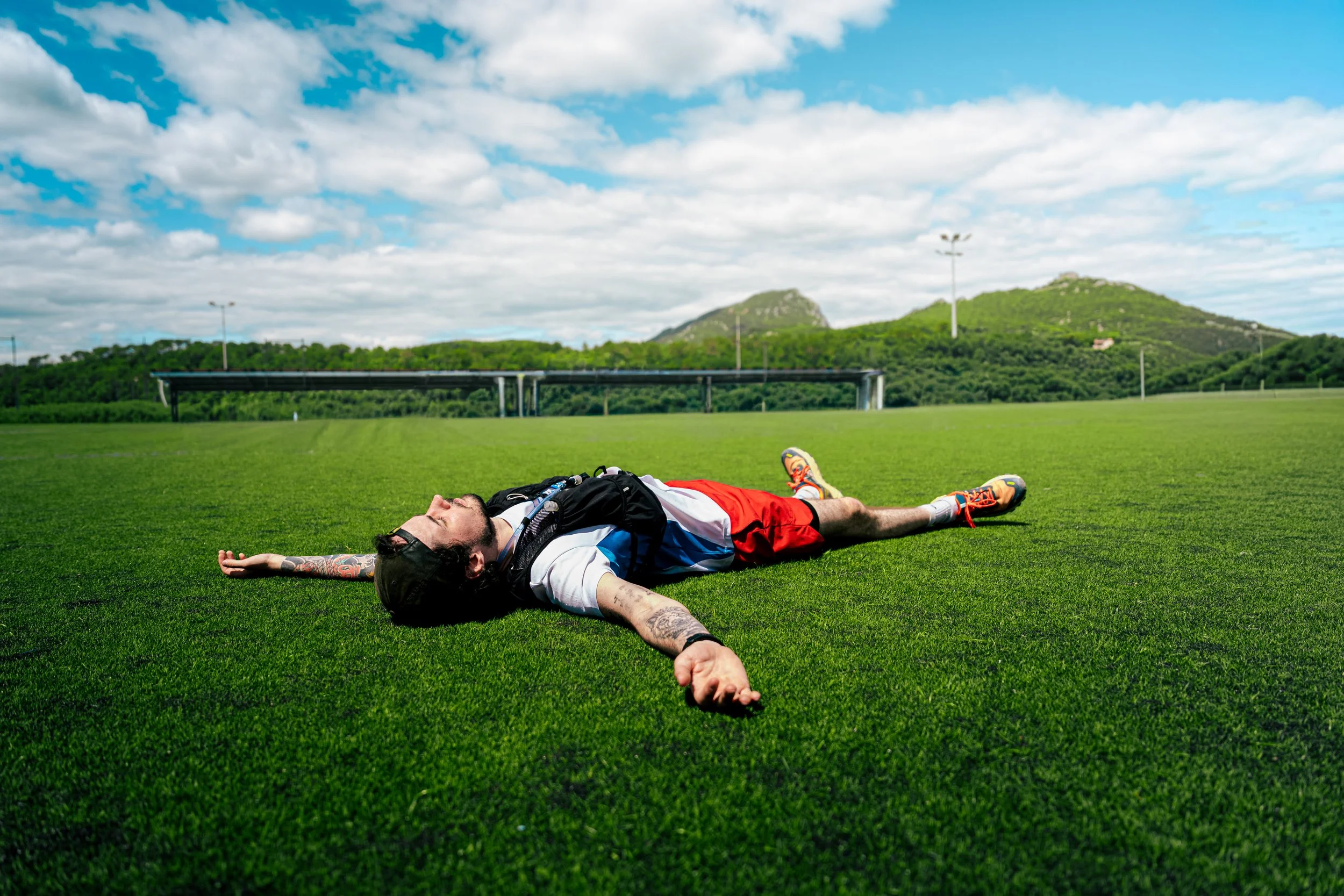 A man laying on the grass with arms and eyes closed, wearing sunglasses, a white T-shirt, red shorts, and sneakers, in a large outdoor field with green hills, a cloudy sky, and some light poles and structures in the background.