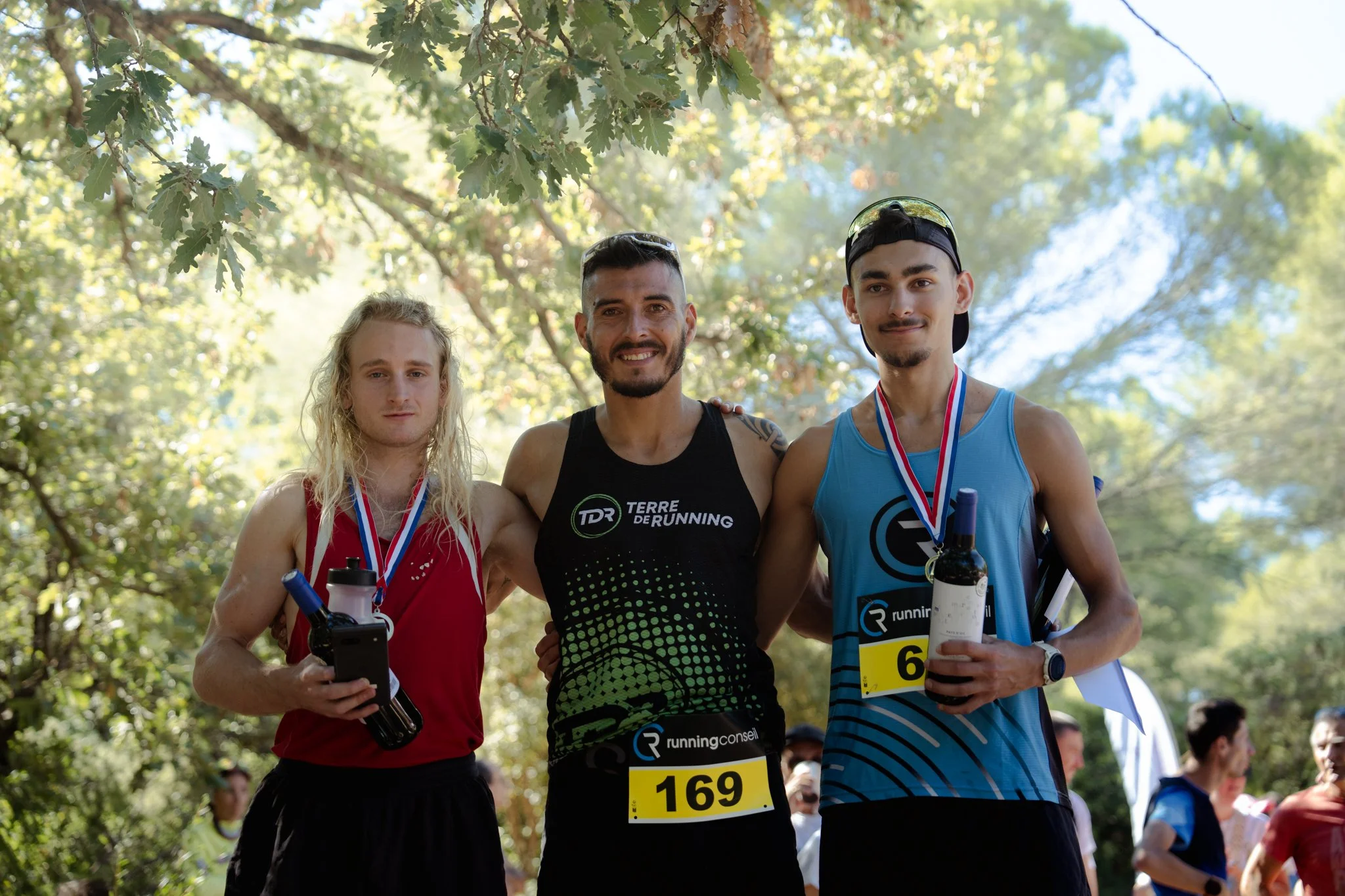Trois jeunes hommes souriants posent ensemble après une course dans un parc. Ils portent des dossards de course, certains avec des médailles autour du cou, et semblent heureux et fiers de leur performance.