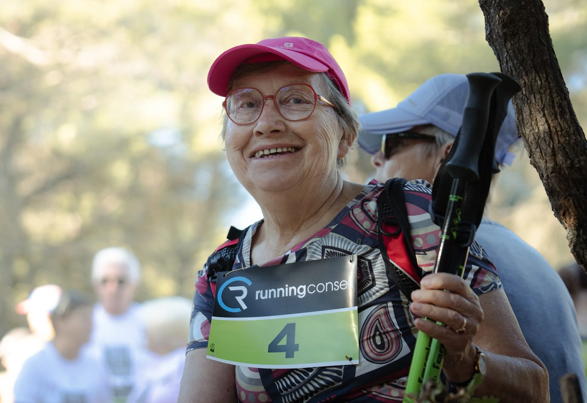 Une femme âgée souriante portant une casquette rose, des lunettes rouges, un t-shirt multicolore, participant à une course ou randonnée en plein air, avec un number bib marqué 4, tenant une canne de marche, et d'autres coureurs en arrière-plan.