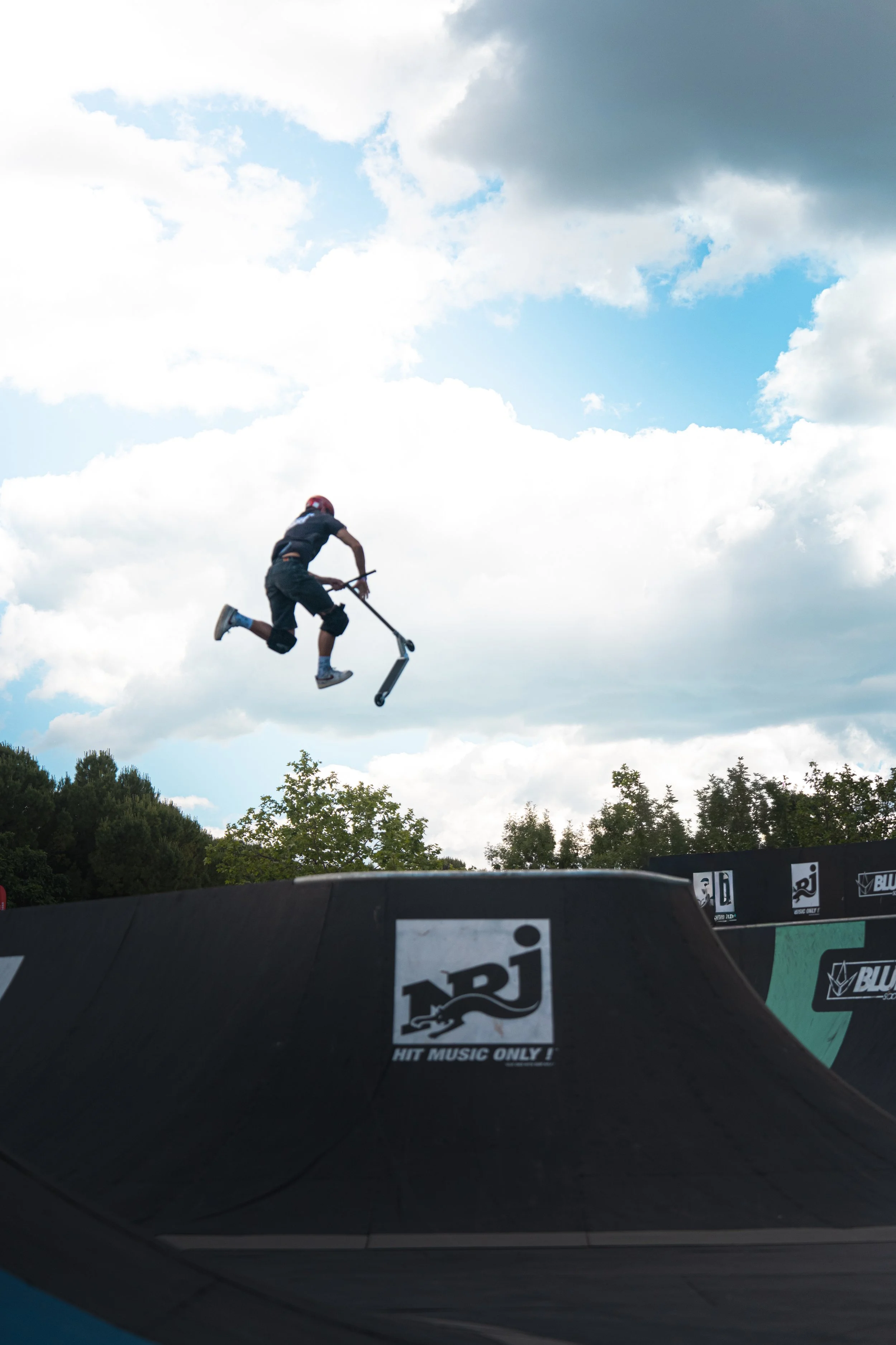 Un jeune homme effectue un saut spectaculaire avec une trottinette lors d'un événement de sport extrême, avec un ciel nuageux en arrière-plan et des barrières publicitaires au premier plan.