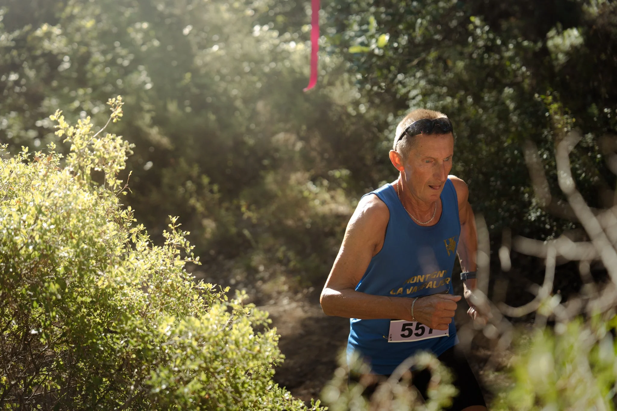 Un homme courant lors d'une course en plein air à travers des buissons et des arbres, portant un t-shirt bleu, des lunettes de soleil sur la tête et un dossard numéro 55.