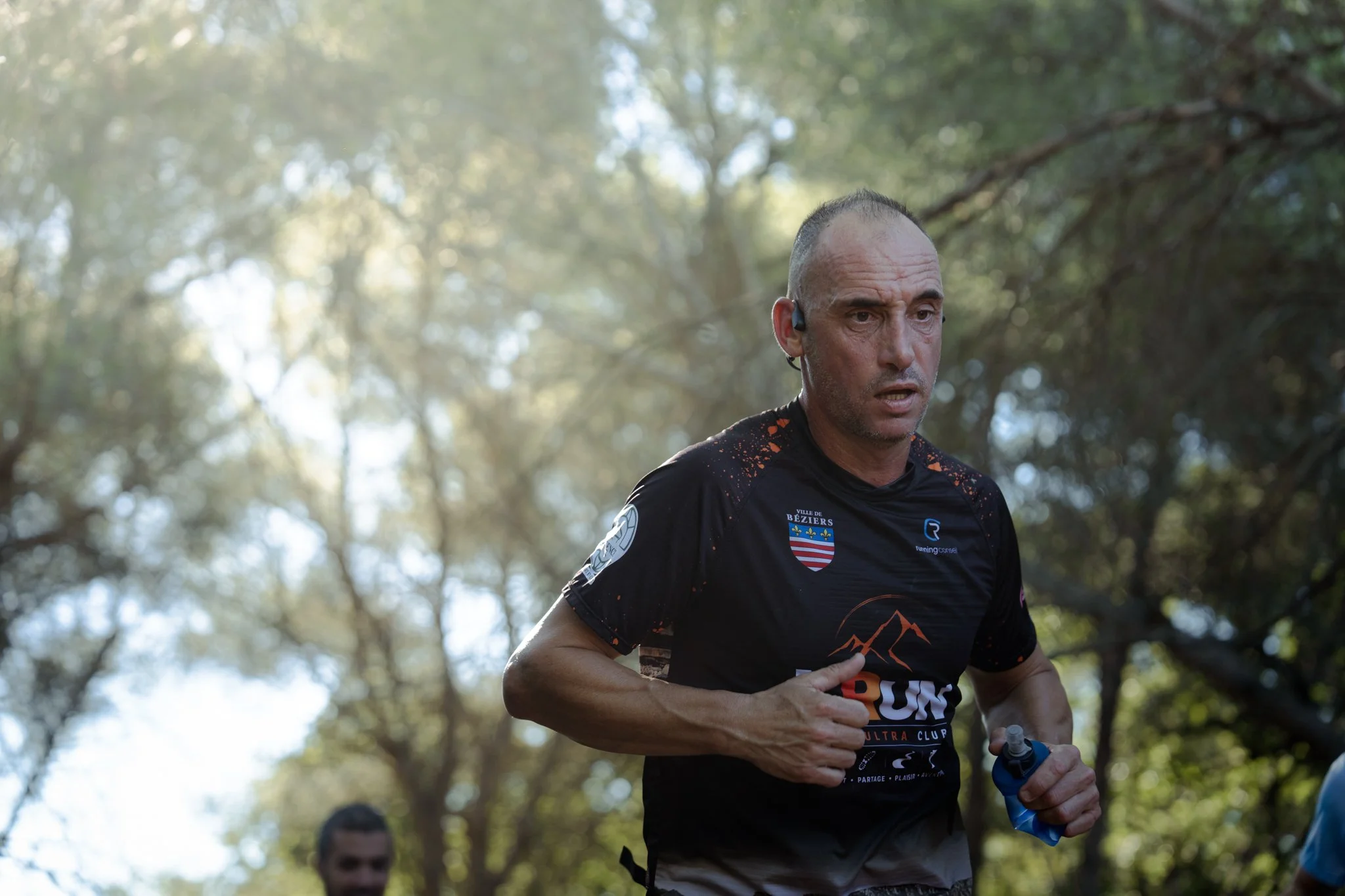Un homme courant dans la forêt, portant un t-shirt de course avec des logos et une puce de chronométrage, lors d'une course ou d'un marathon.