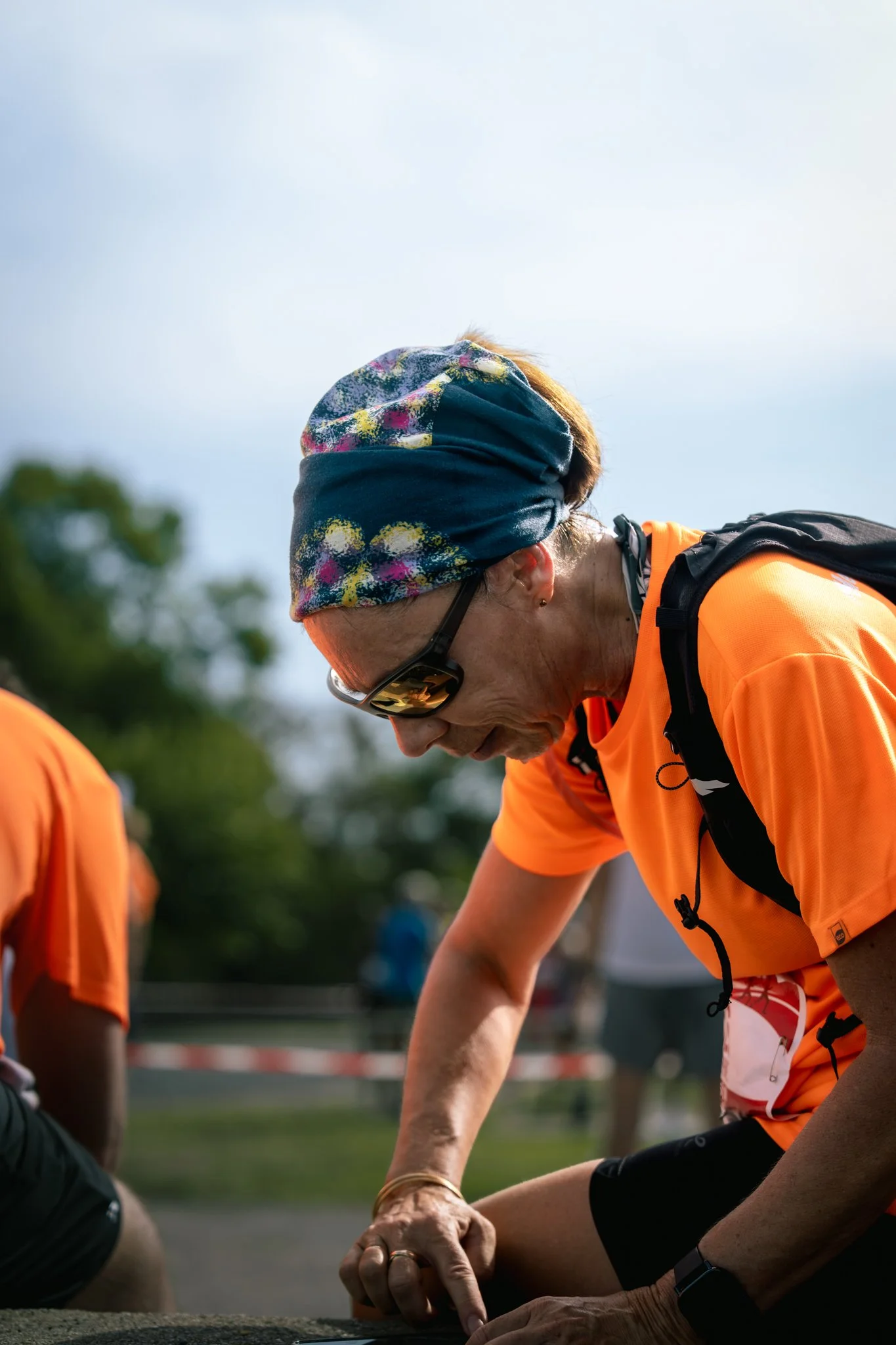 Une femme portant un bandeau coloré, des lunettes de soleil et un t-shirt orange participe à une activité sportive à l'extérieur.