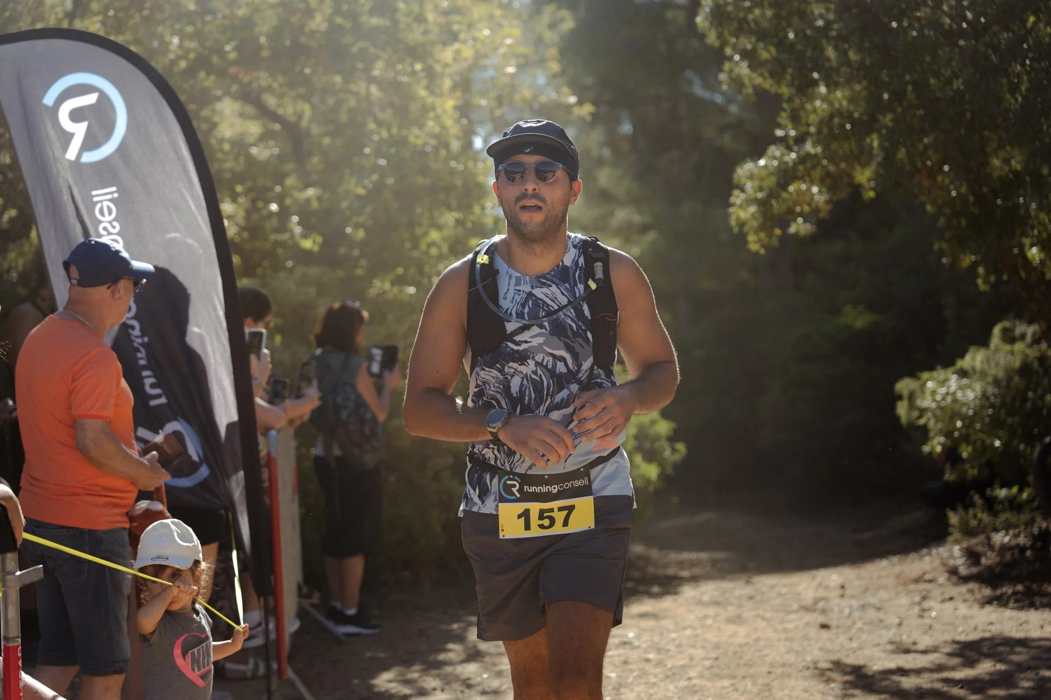 Un homme court lors d'une course dans un espace naturel, portant un t-shirt à motifs, des lunettes de soleil, un casque et un accessoire de course, avec plusieurs spectateurs en arrière-plan.