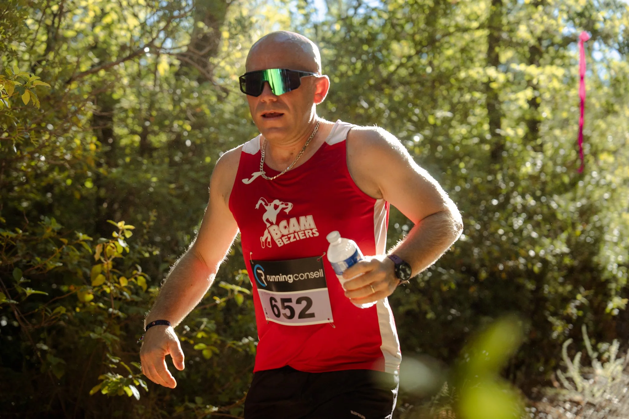 Un homme participe à une course en forêt, portant un maillot rouge avec le logo 'Bcam Beziers' et un numéro de dossard 652. Il porte des lunettes de soleil, une montre, et tient une bouteille d'eau.