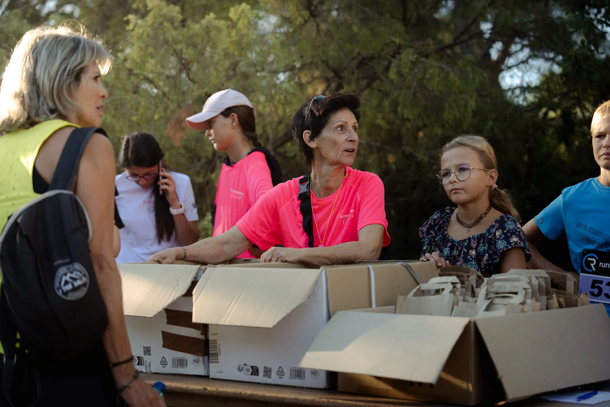 Groupe de personnes, principalement des femmes, autour d'une table en carton dans un parc, discutant et travaillant ensemble, avec des arbres en arrière-plan.