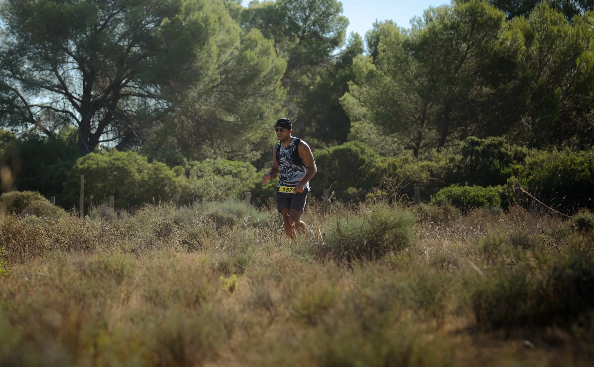 Un homme court dans une prairie entourée d'arbres, portant un t-shirt, un short, des lunettes de soleil et un casque.