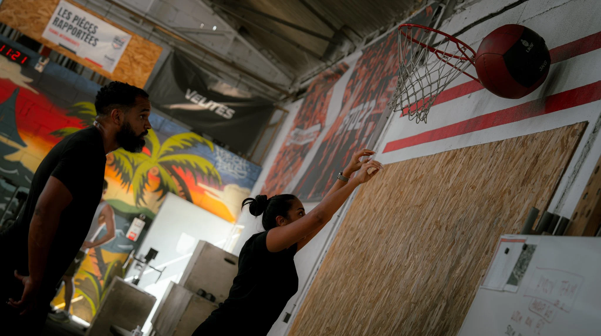 Deux personnes jouent au basketball dans une salle d'entraînement. Une femme est en train de tirer au panier, tandis qu'un homme observe. Le mur en arrière est décoré avec des peintures colorées et il y a un panneau avec le texte "Les pièces rapporté