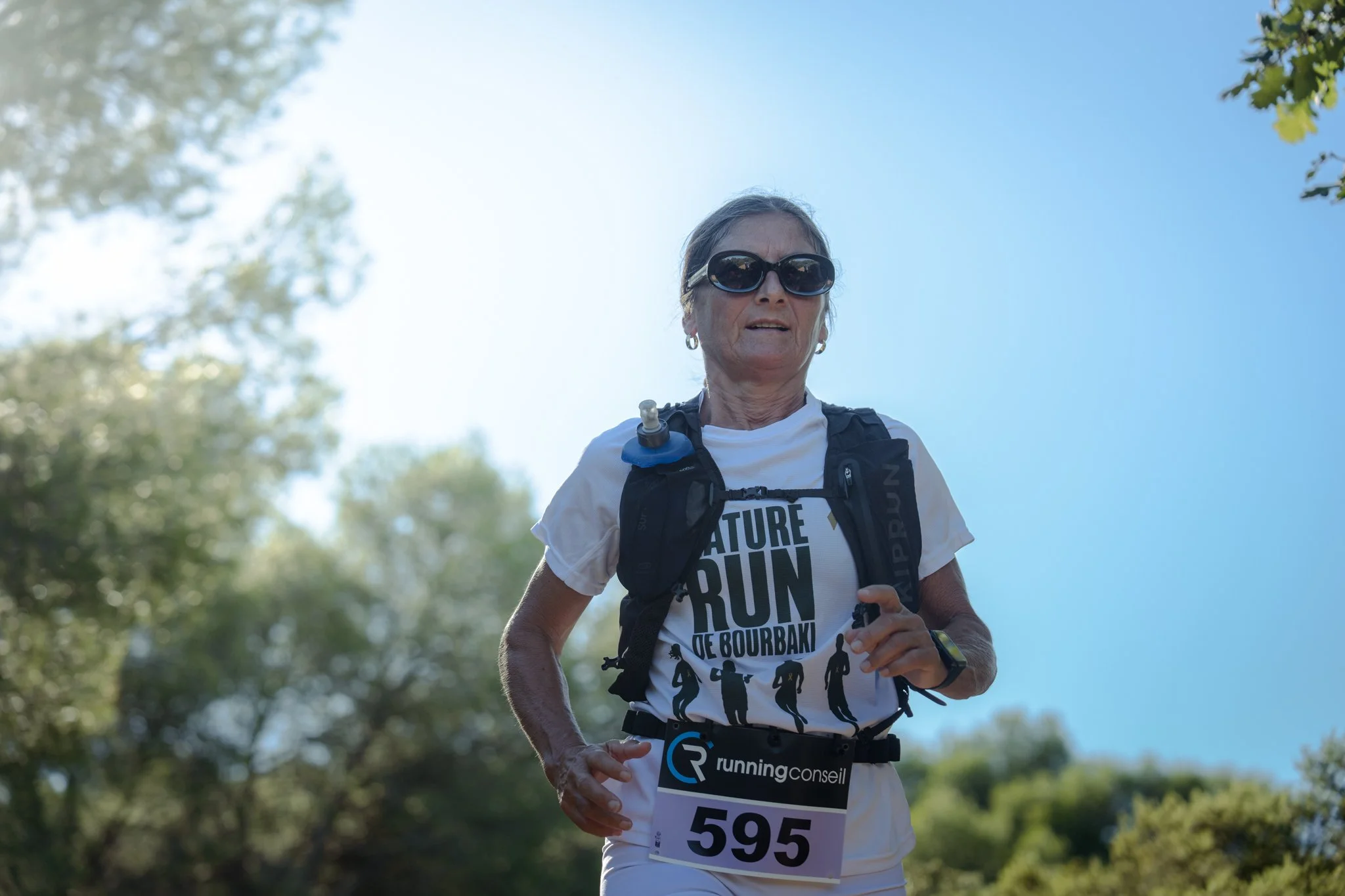 Une femme âgée court en pleine nature avec des lunettes de soleil, portant un t-shirt blanc avec un message sur le marathon et un dossard, dans un environnement en plein air ensoleillé.