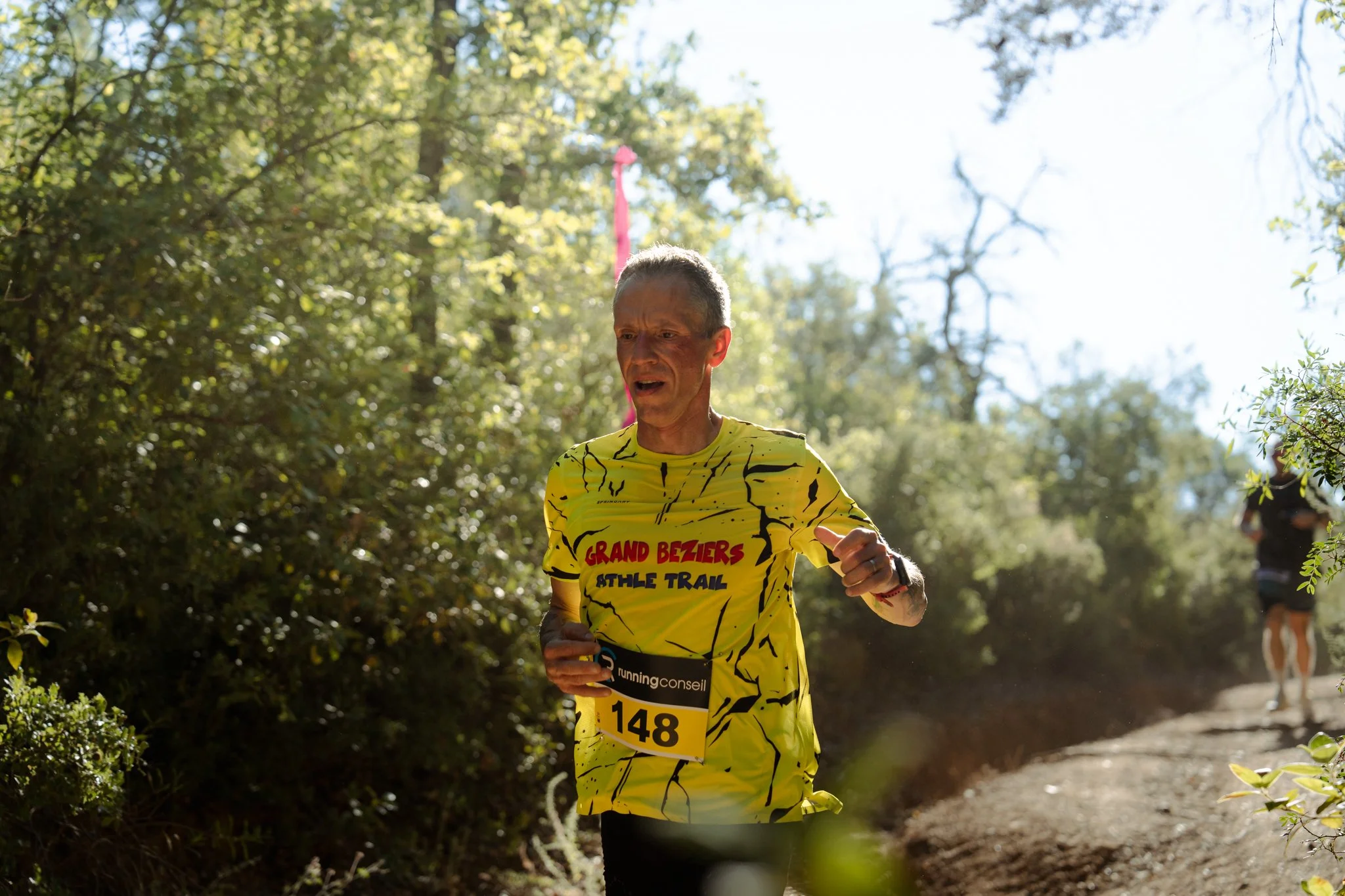 Un homme court dans un trail en forêt sous un ciel ensoleillé, portant un maillot jaune avec des inscriptions "Grand Baziers Athle Trail" et le numéro 148.