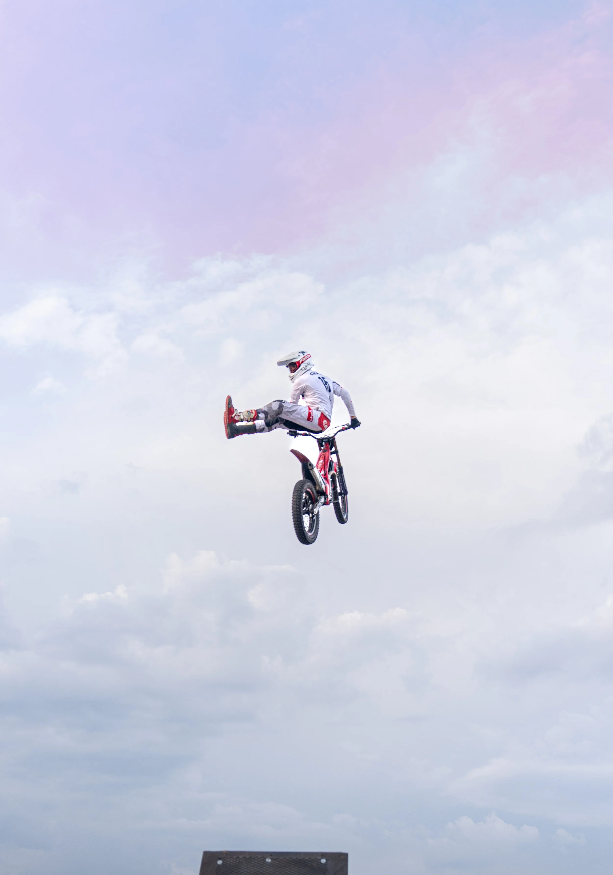A motocross rider in white gear and helmet performing a mid-air stunt on a dirt bike against a cloudy sky.