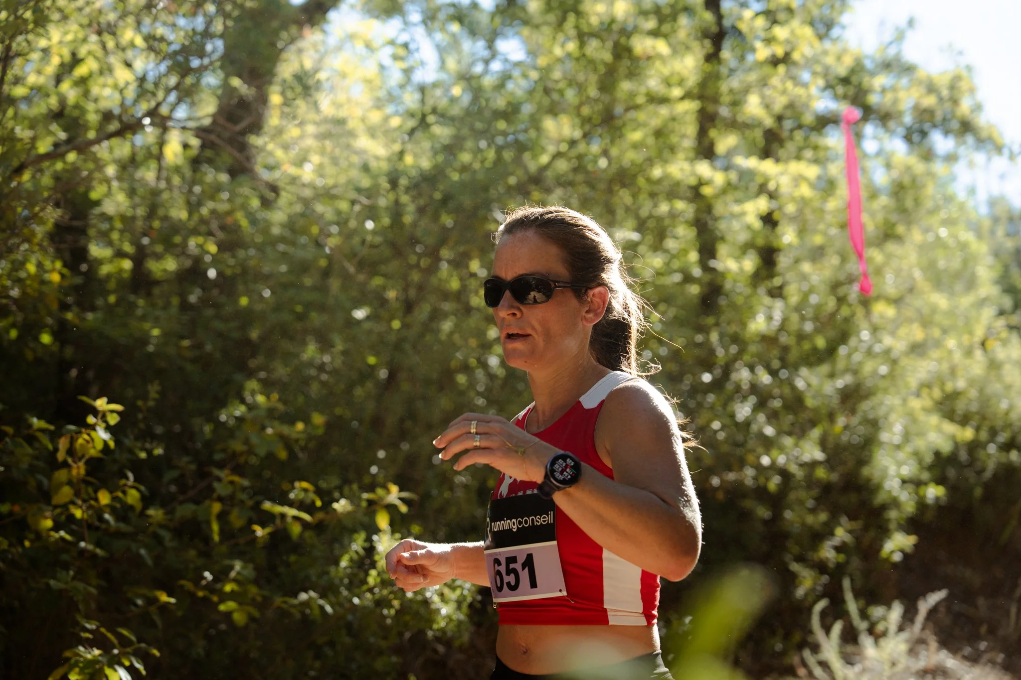 Femme courant dans un environnement forestier avec des arbres verts, portant des lunettes de soleil, un haut de course rouge et une montre connectée, numéro de course 651.