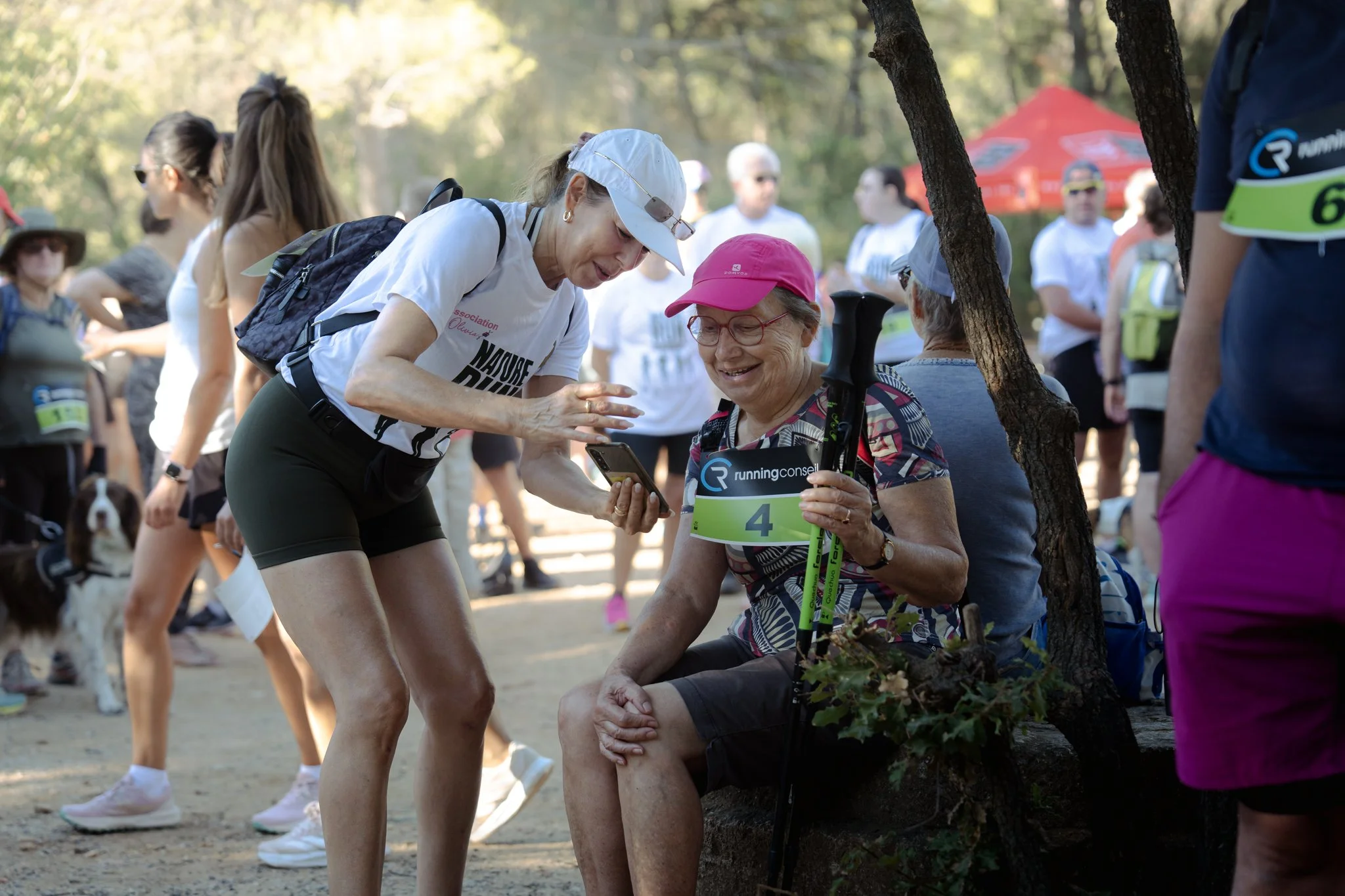 Capture d'une course en plein air avec plusieurs coureurs, dont une femme aidant une autre femme âgée avec une canne, sous des arbres, lors d'un événement sportif.